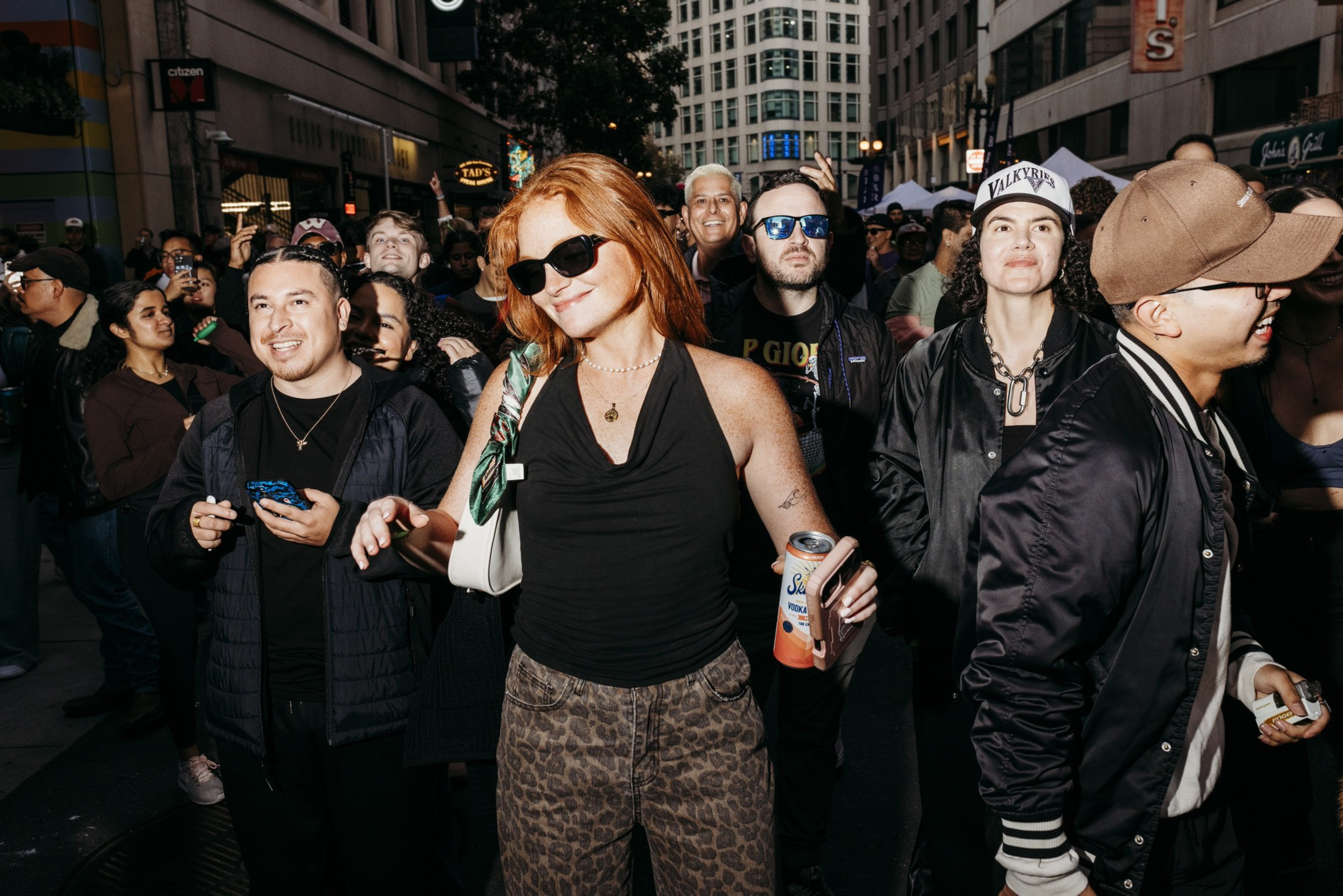 A group of diverse people stand in a crowded urban street at night, some smiling and wearing casual clothes and sunglasses, enjoying a lively event.