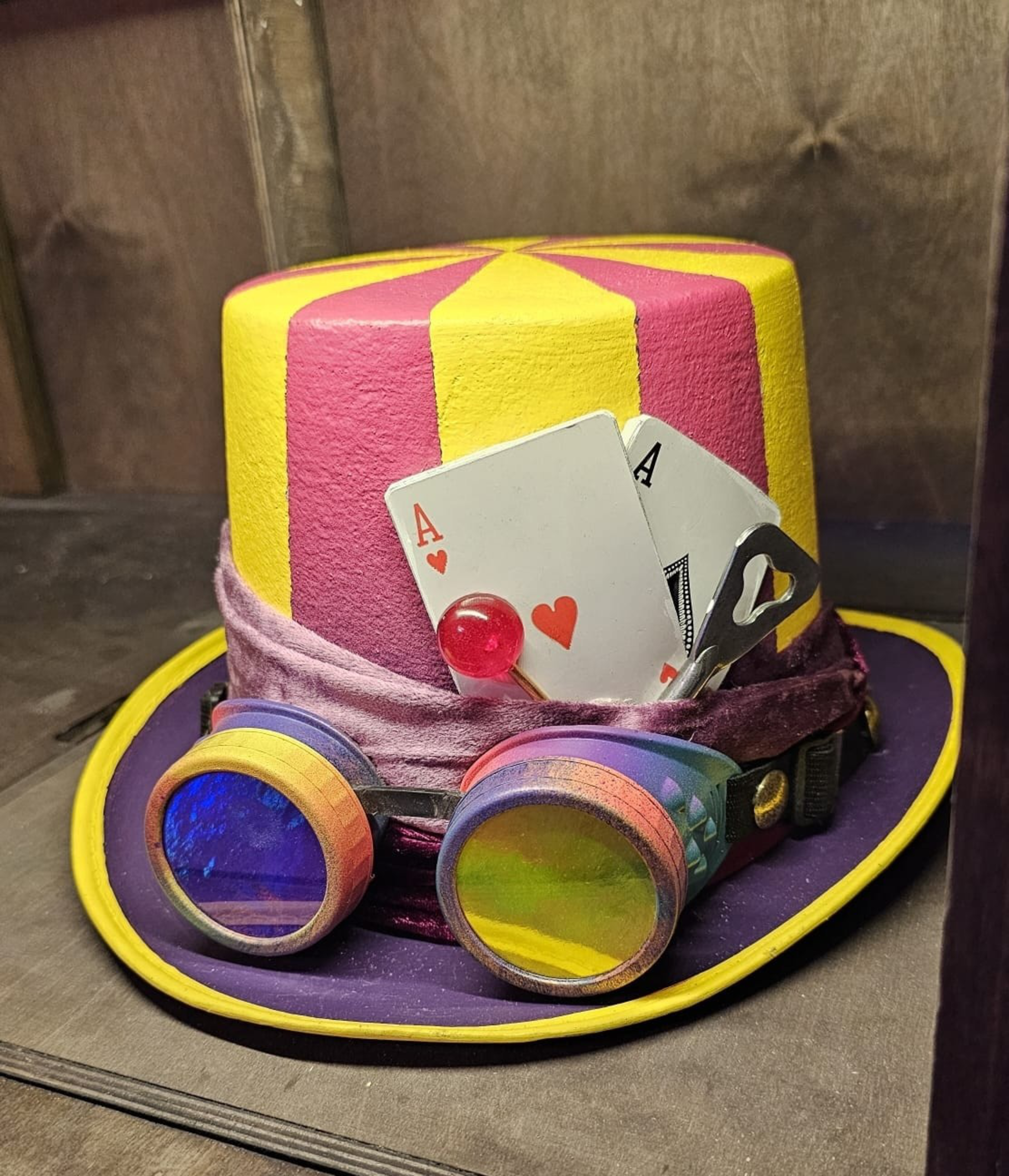 A vibrant top hat features yellow and pink stripes, adorned with playing cards and a lollipop. Goggles rest at its base, and it’s wrapped with a purple band.
