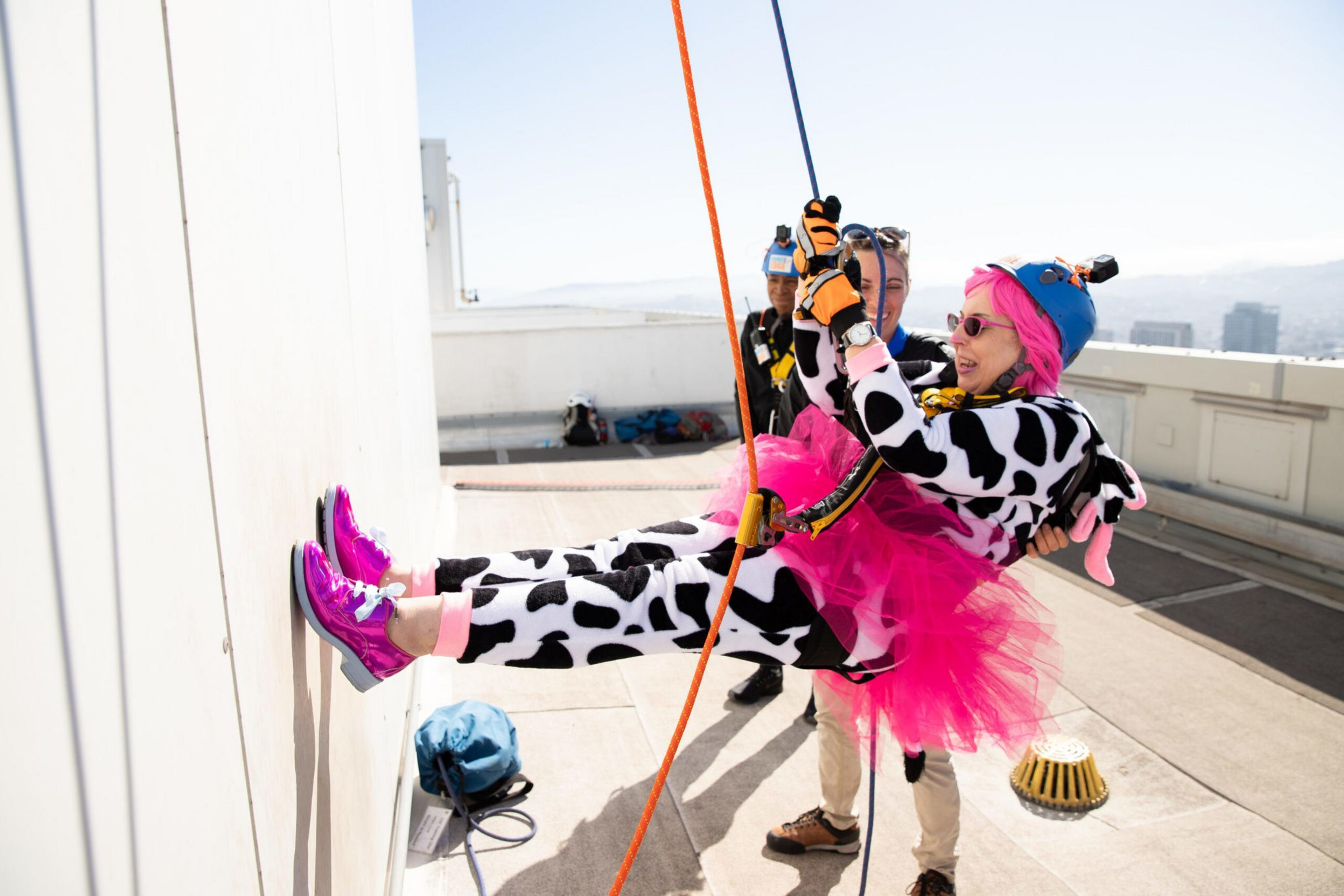 A person in a cow onesie, pink tutu, and helmet with a camera is rappelling down a wall, supported by two others on a rooftop.