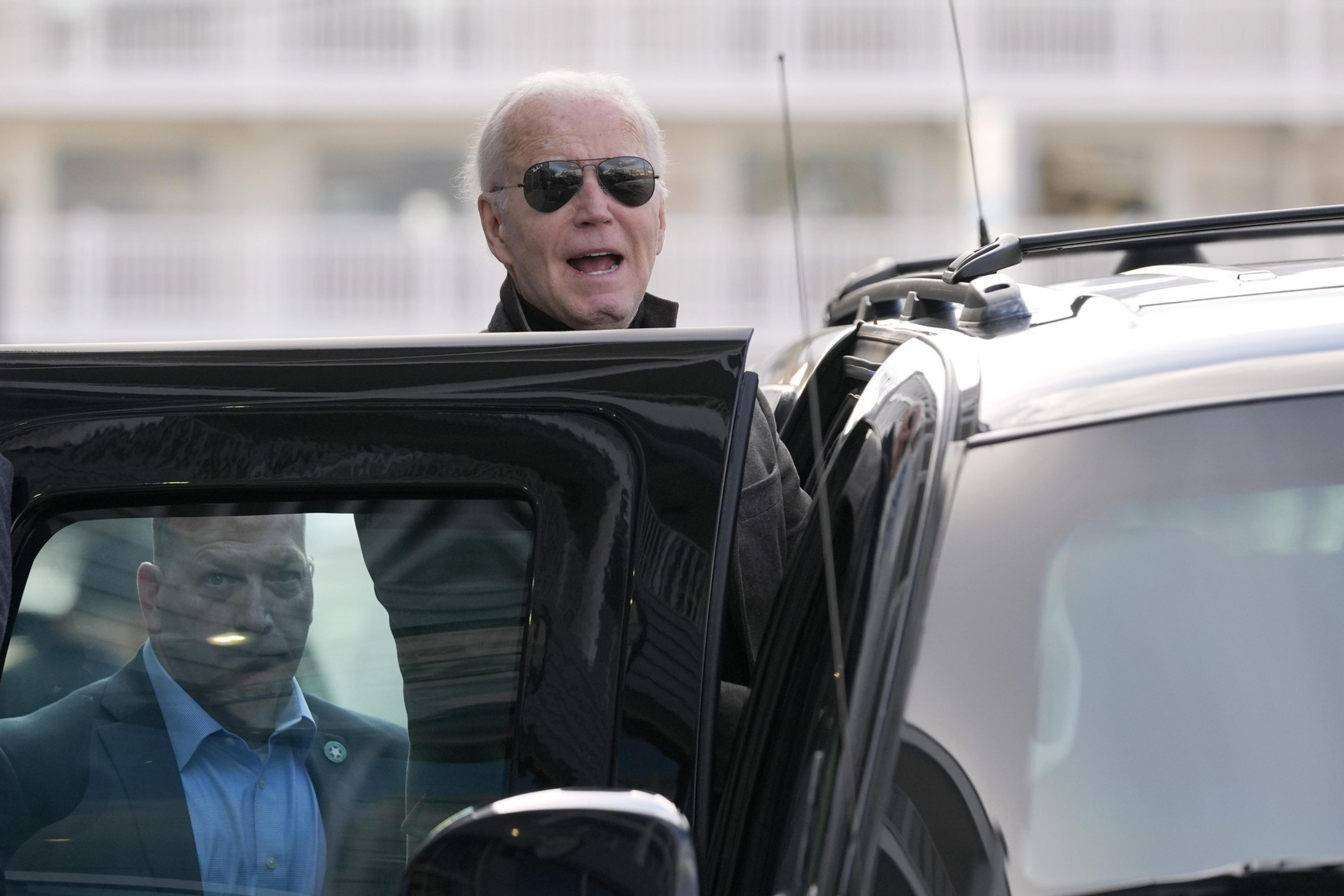 A person in sunglasses is exiting a car, with another person reflected on the vehicle's window.