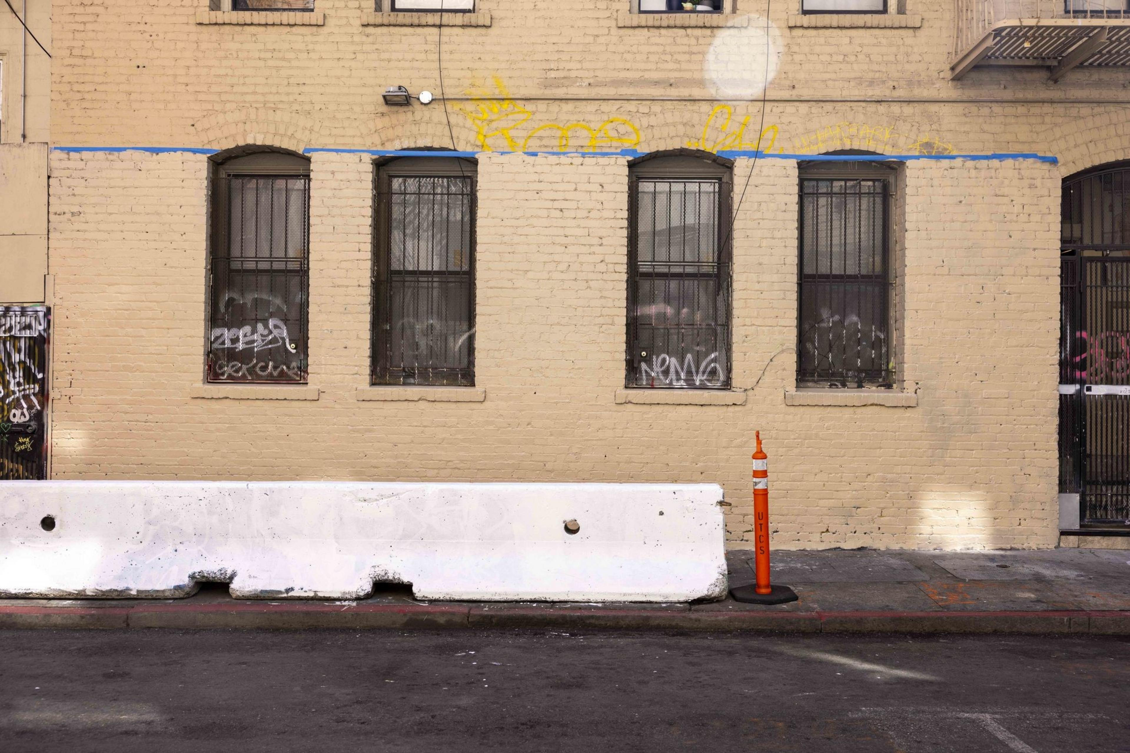 A beige brick wall with barred windows covered in graffiti has a white barricade and orange cone, labeled "UTCS," in front on the street.