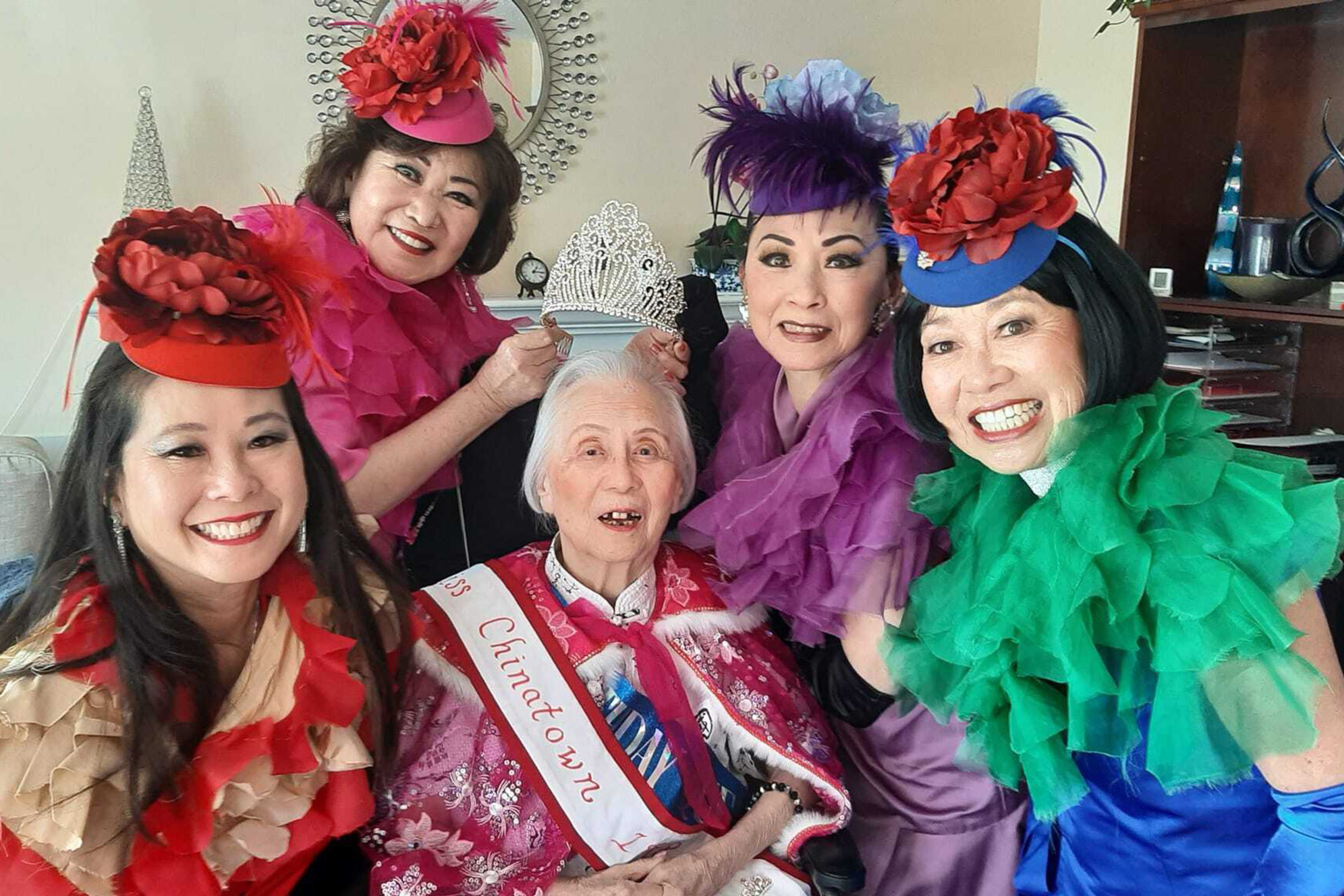 Five women dressed in fancy clothes crowd together around a woman wearing a Miss Chinatown banner.