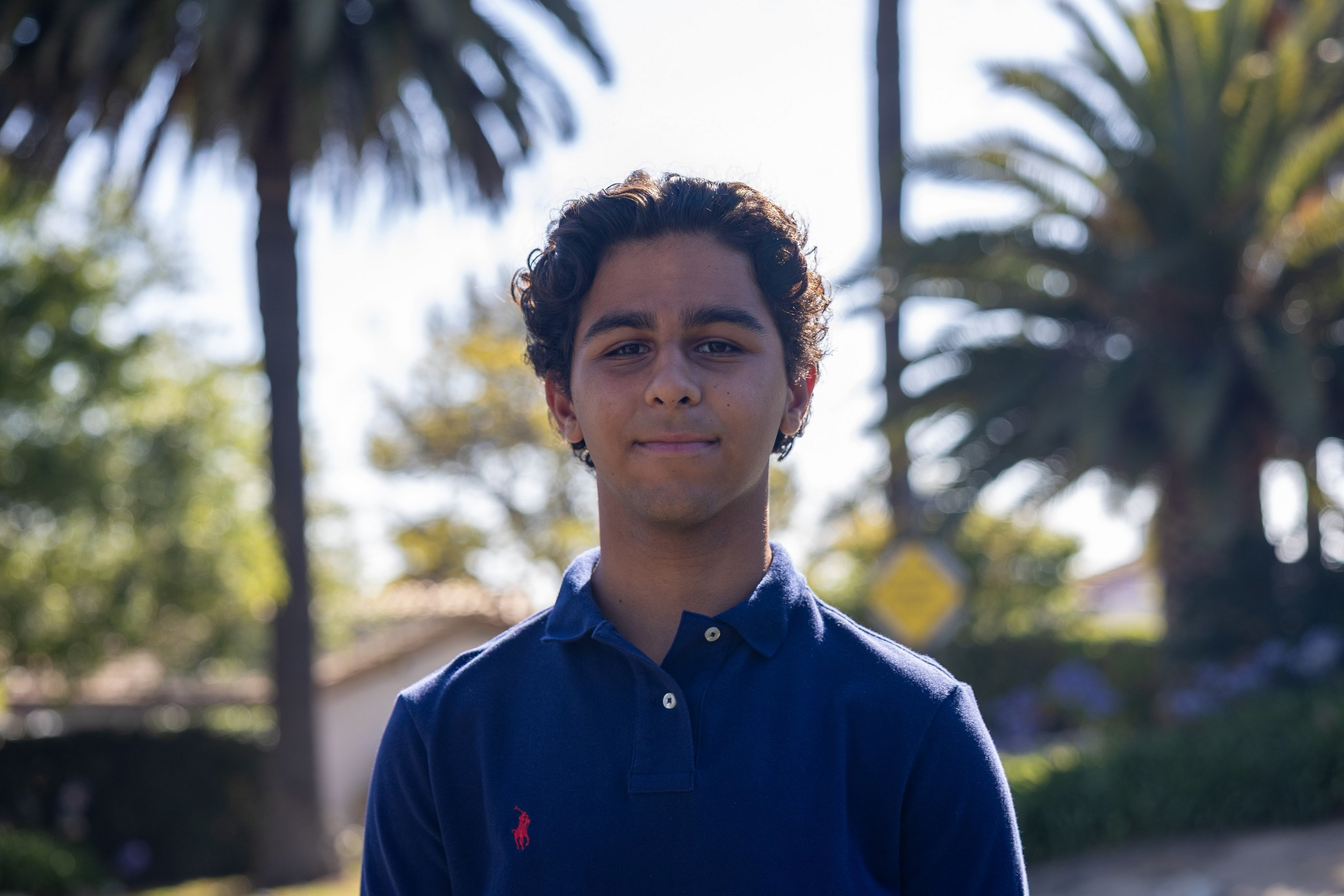 A young man with curly dark hair wears a navy blue polo shirt and stands outdoors with blurred palm trees and greenery in the background.