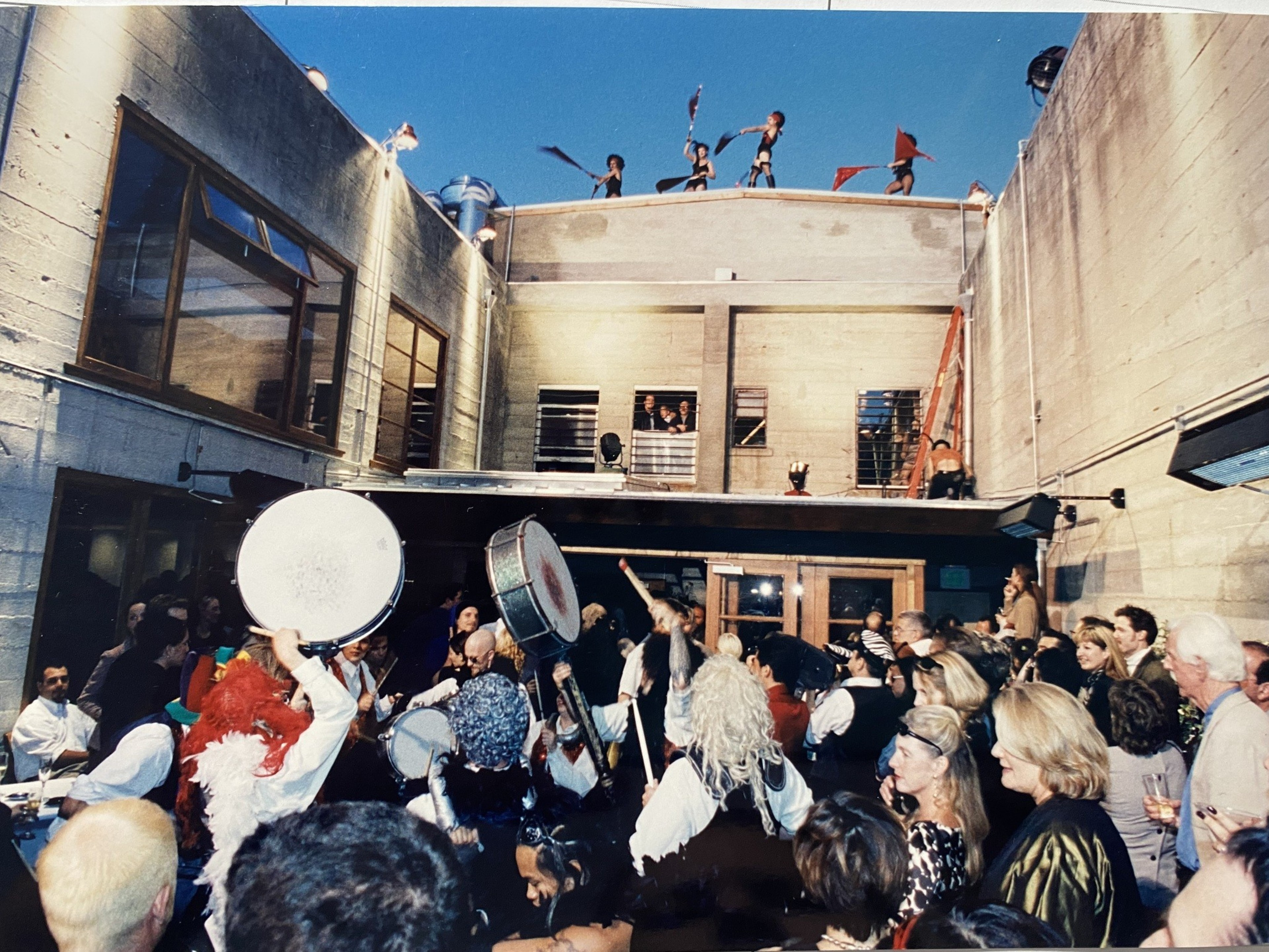 A lively party with a large crowd in a courtyard, featuring costumed drummers and performers on a rooftop waving flags, creating a festive atmosphere.