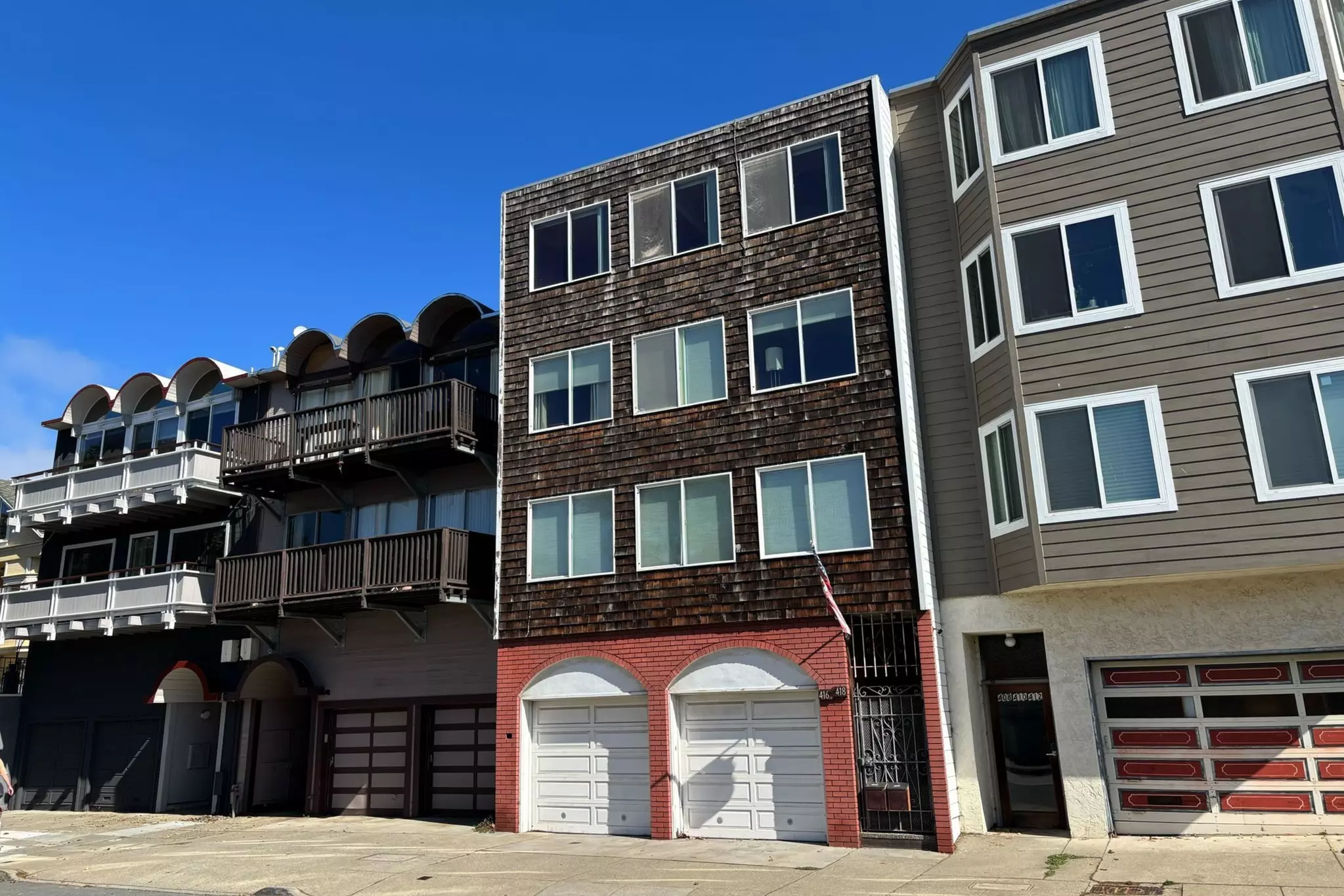 Three multistory residential buildings stand side by side; the middle one has wooden shingles and two white garage doors at street level.