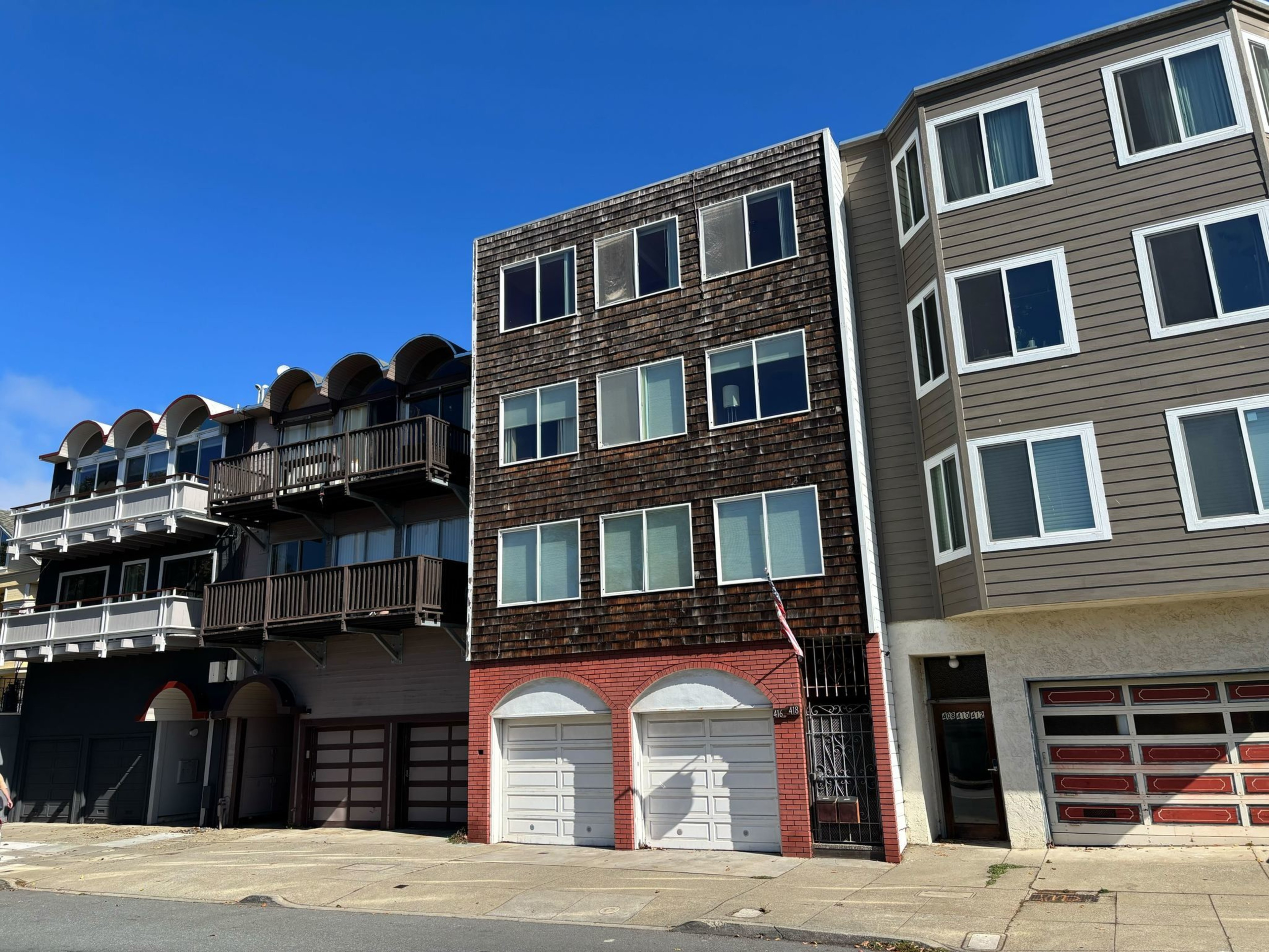 Three multistory residential buildings stand side by side; the middle one has wooden shingles and two white garage doors at street level.