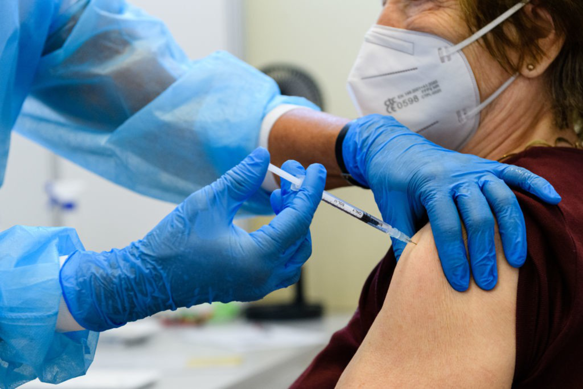 A healthcare worker wearing blue gloves and protective clothing administers a vaccine to an elderly person wearing a white face mask.