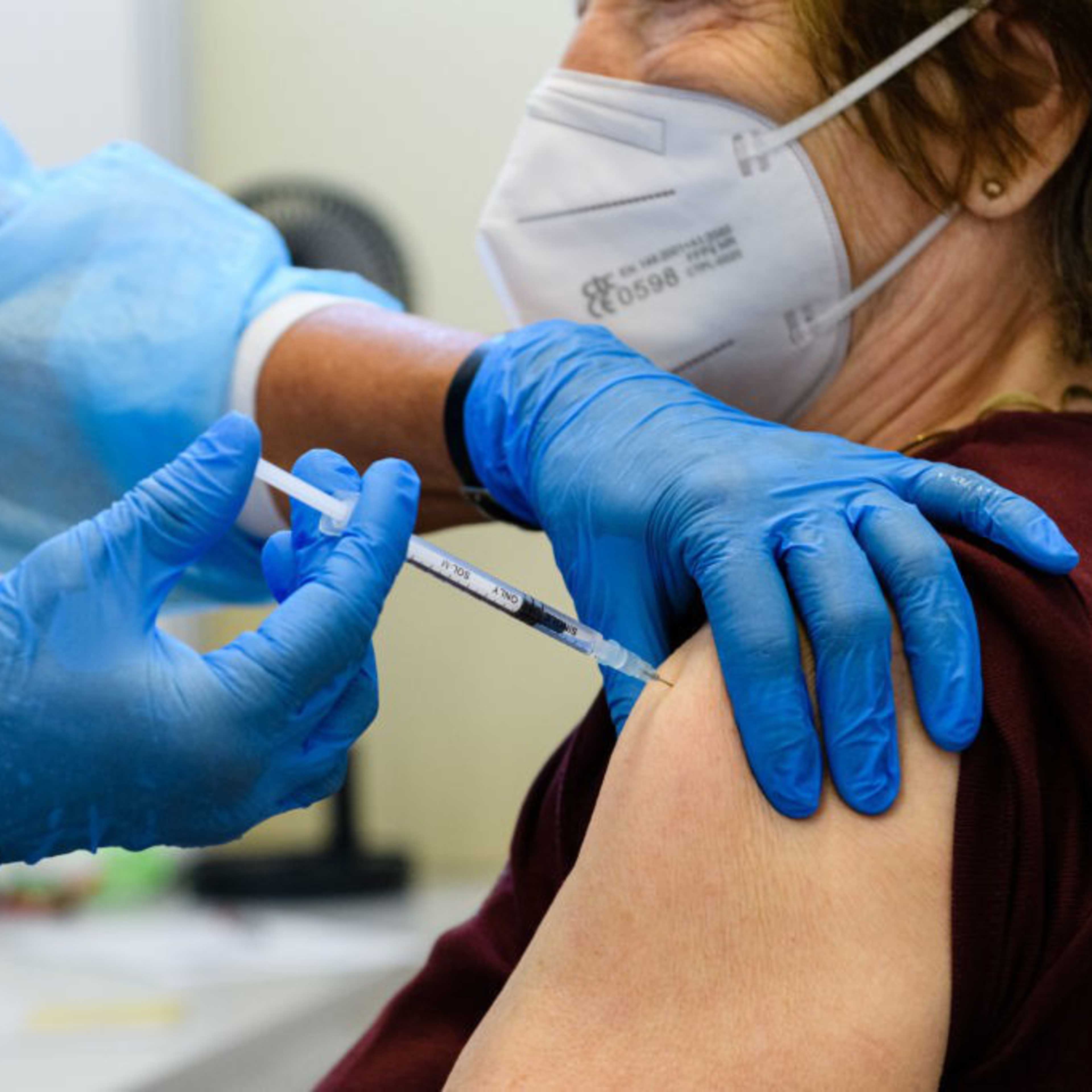 A healthcare worker wearing blue gloves and protective clothing administers a vaccine to an elderly person wearing a white face mask.