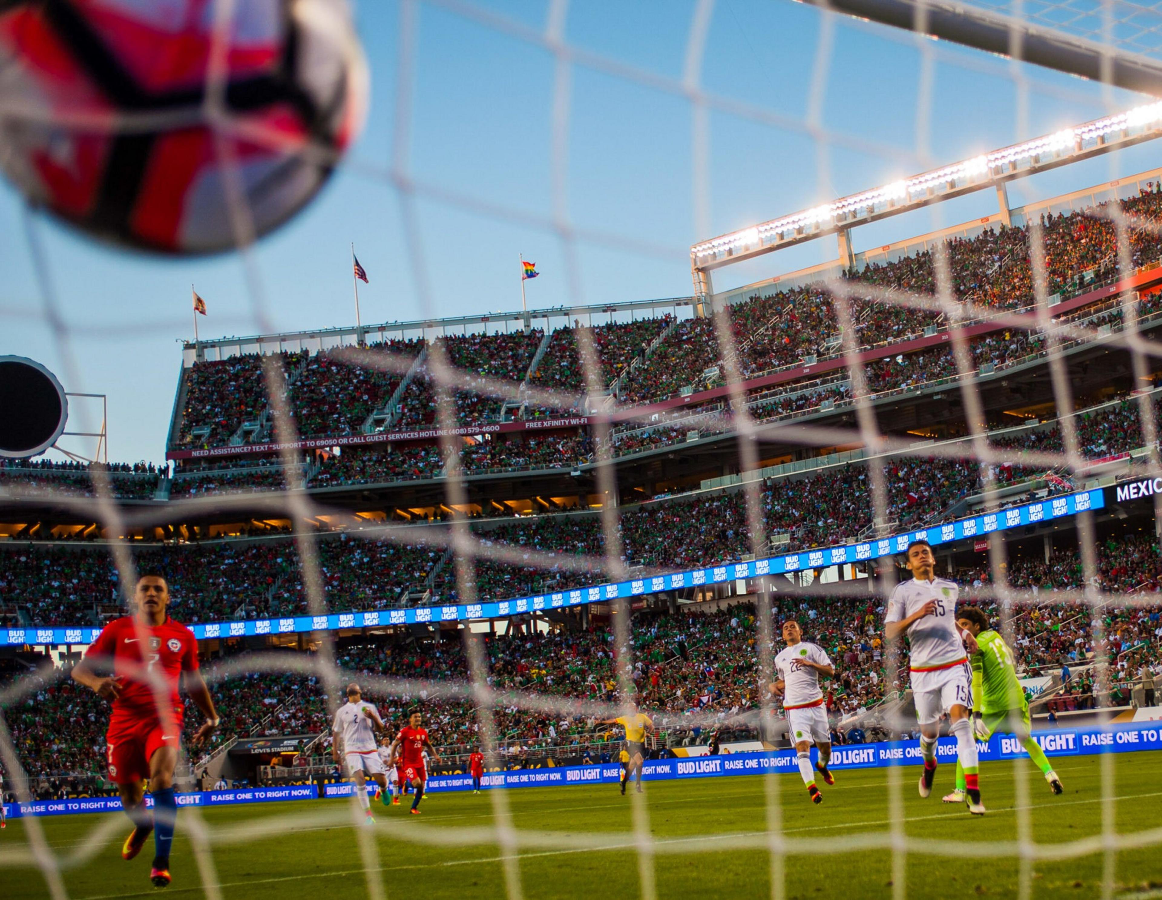 A soccer ball is captured mid-air heading towards the net, with players in red and white jerseys on the field, and a large crowd fills the stadium seats.