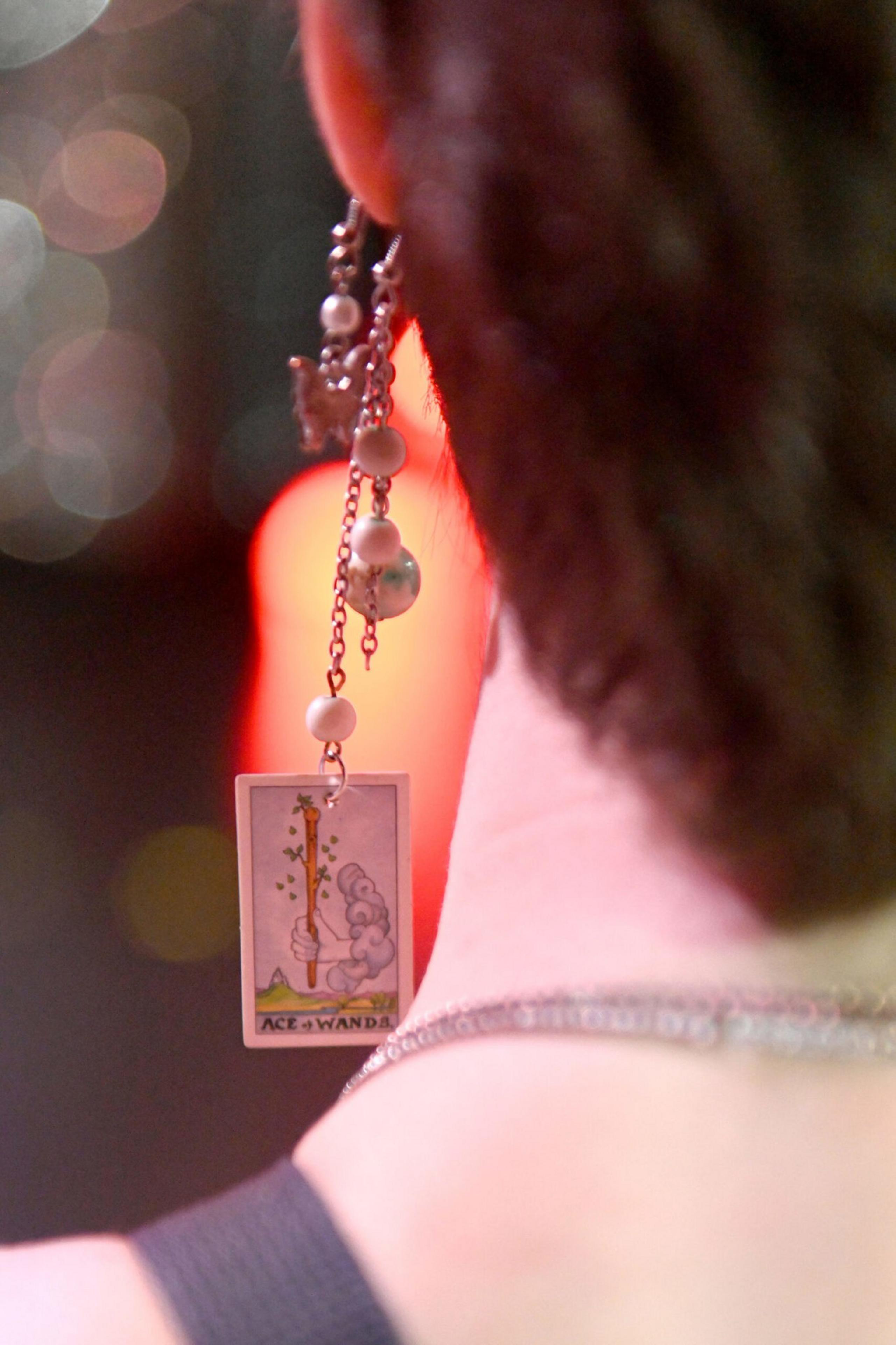 An earring with a small dangling Ace of Wands tarot card, pearls, and chains hangs from a person's ear against a blurred, colorful background.
