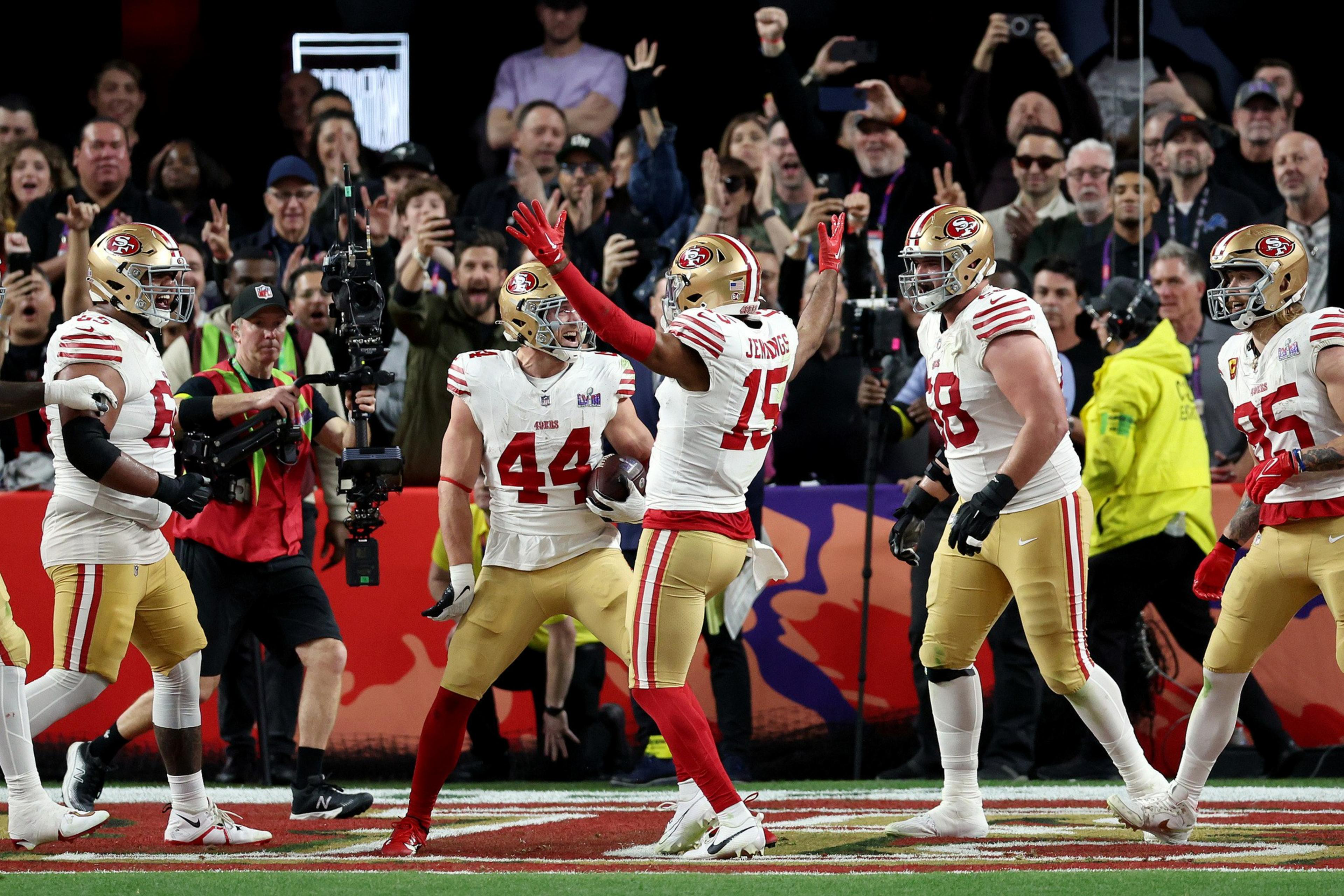 Football players in red and gold uniforms celebrate on the field, surrounded by cheering fans and cameramen capturing the moment.