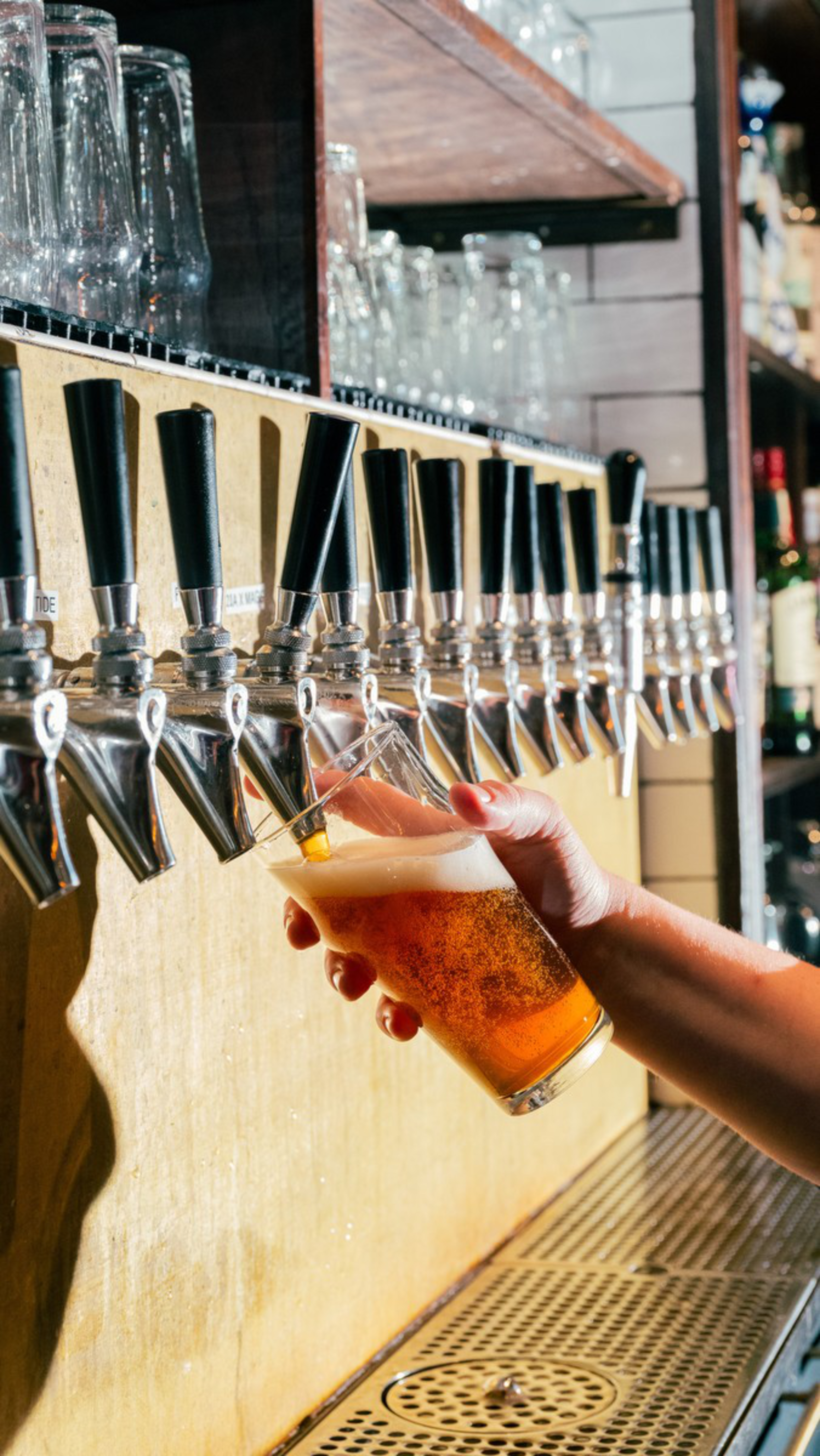 A hand holds a tilted glass filling amber beer with foam from one of many black-handled taps on a bar.