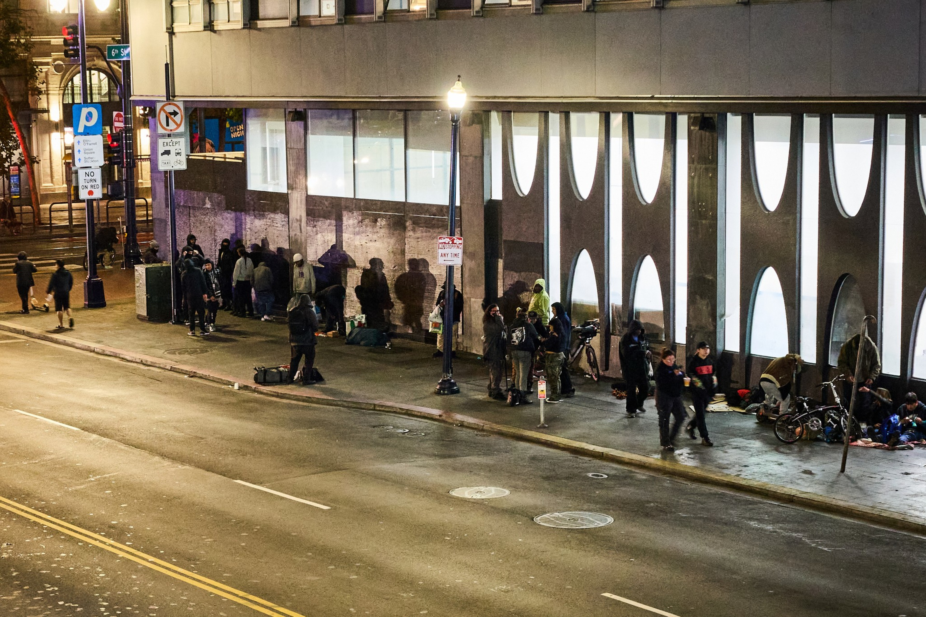 A group of people gather on a city sidewalk at night, near a building with unique oval windows. Streetlights illuminate the area, with signs and bicycles visible.