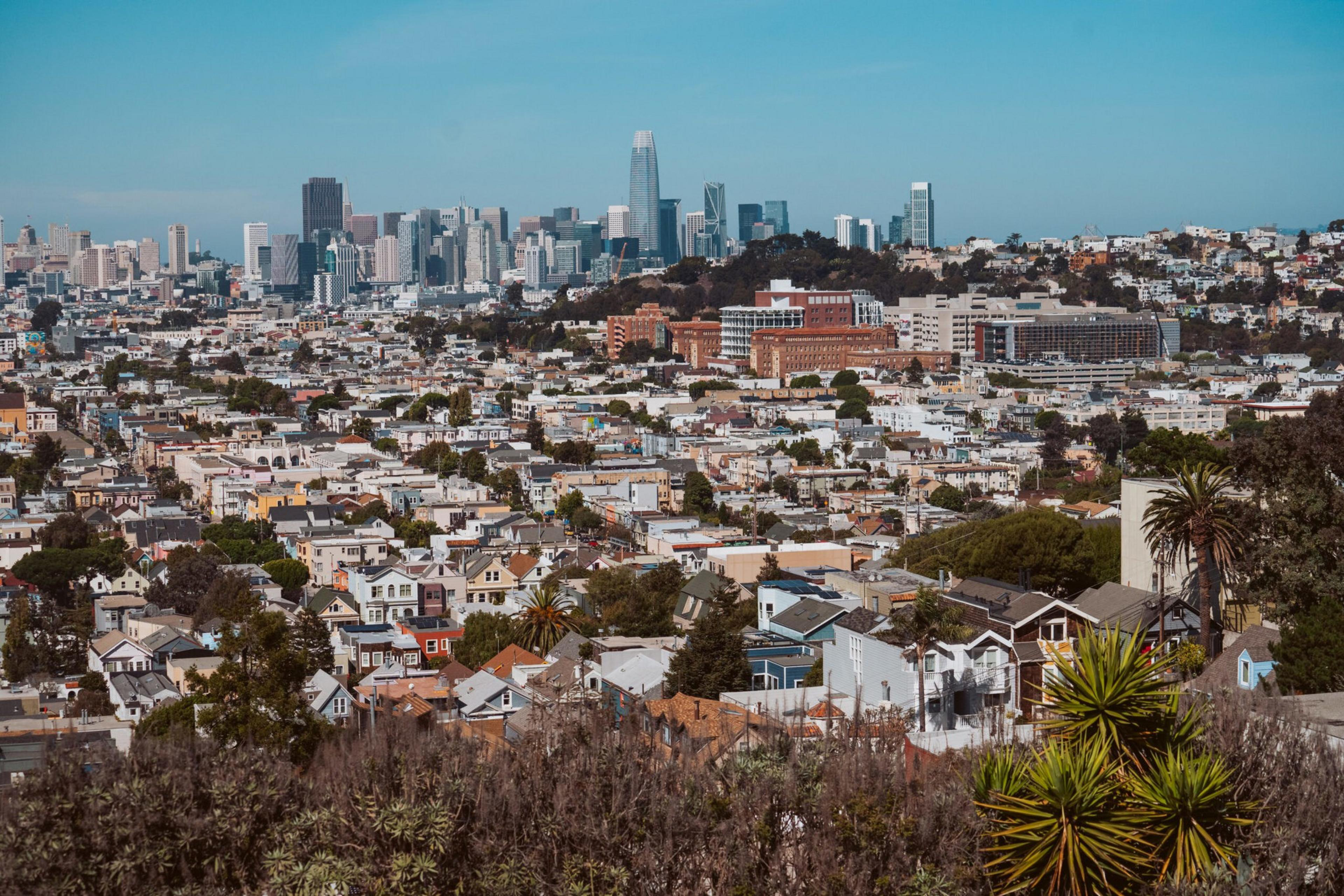 A sprawling urban landscape with densely packed houses in the foreground and a cluster of tall skyscrapers in the background under a clear blue sky.