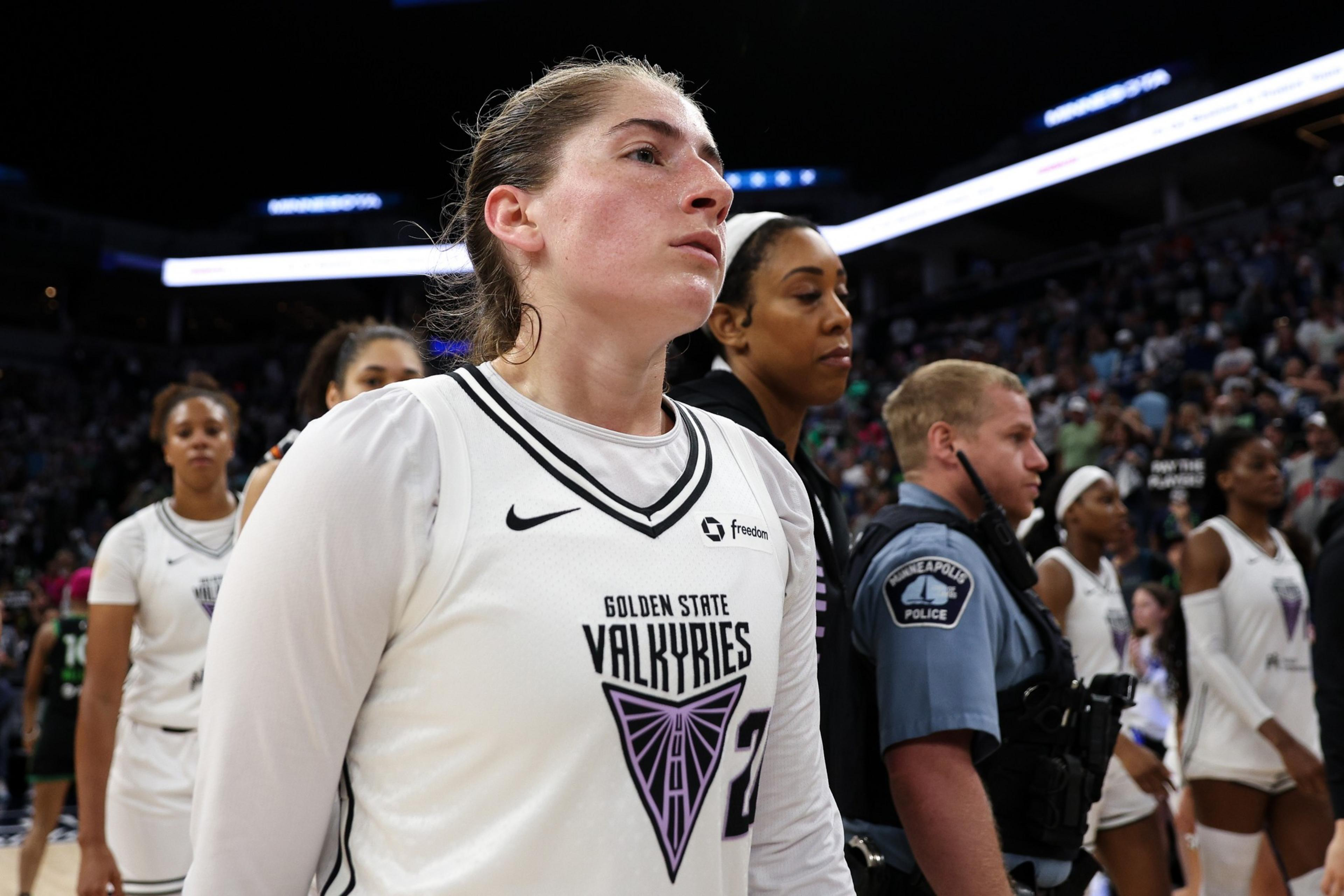 A female basketball player wearing a Golden State Valkyries jersey stands with teammates and a Minneapolis police officer in a crowded indoor arena.