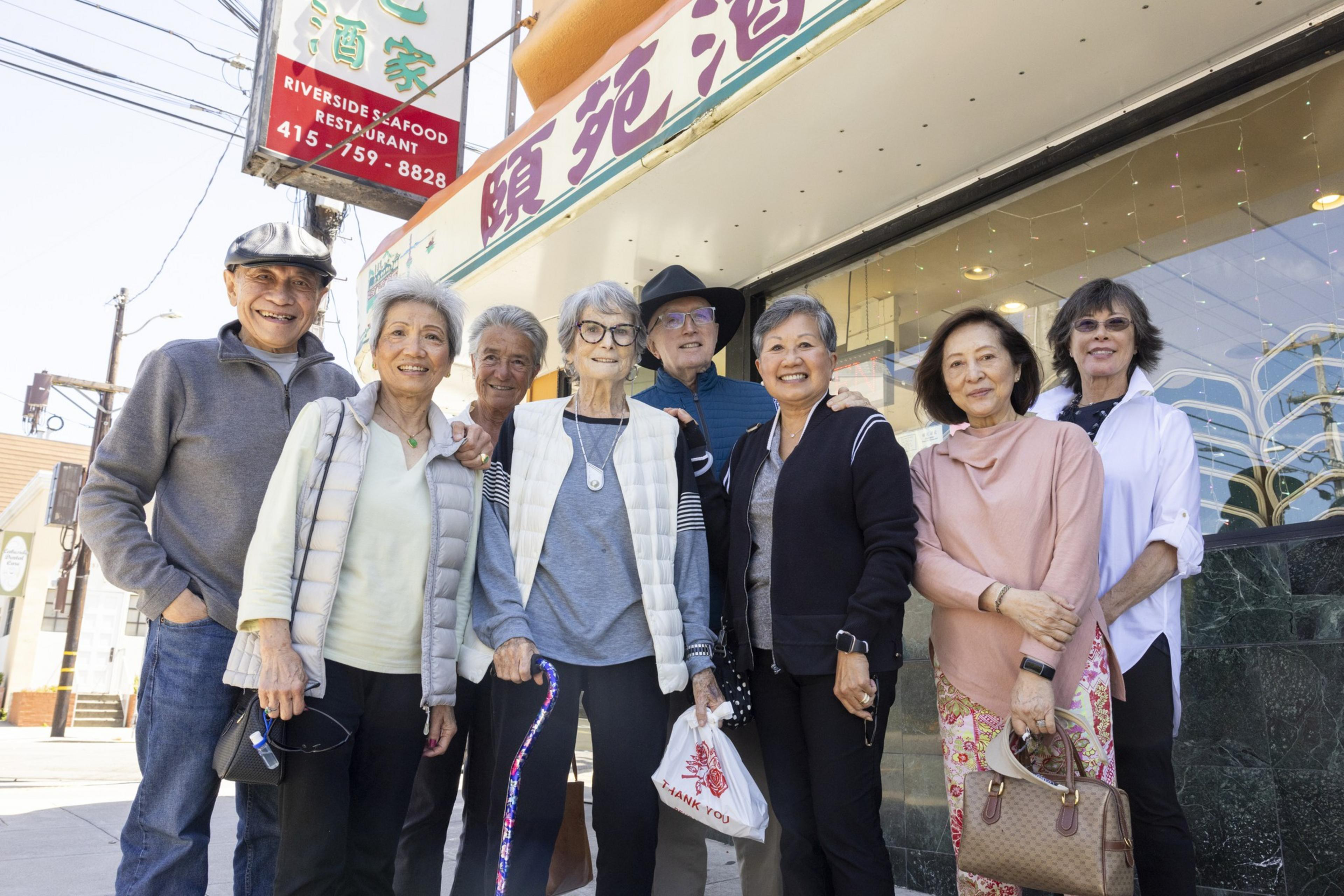 A group of eight smiling elderly individuals stands outside a Riverside Seafood Restaurant, posing for a photo. They seem happy and sociable, dressed casually.
