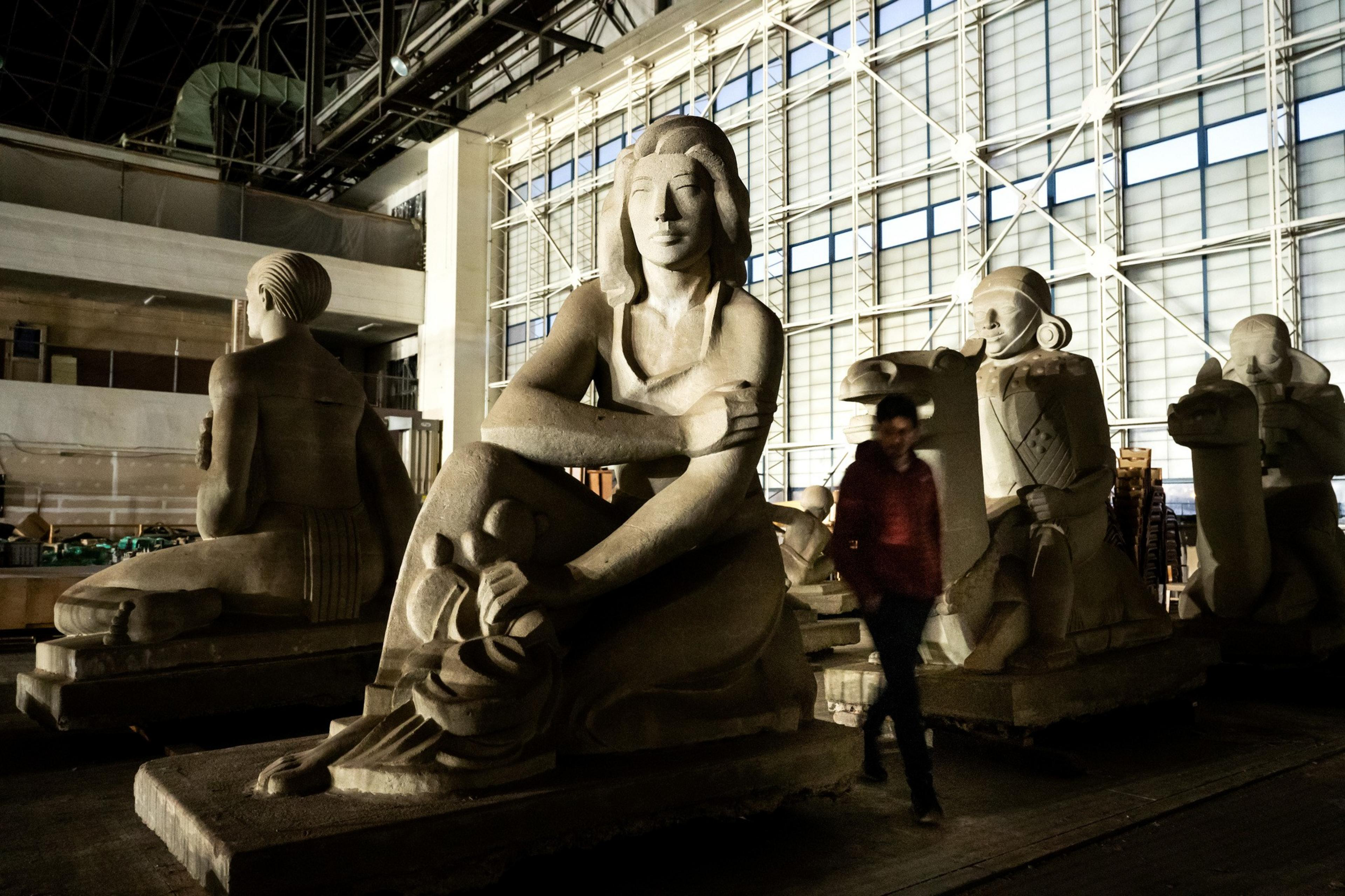 The image shows large stone sculptures of seated figures in a dimly lit, spacious room with a high ceiling and grid-like window patterns. A person walks by.