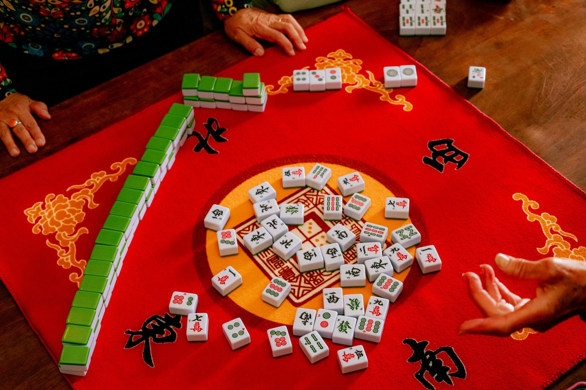 A red cloth with black Chinese characters holds scattered Mahjong tiles, a long row of green-backed tiles, and two hands near the table's edges.
