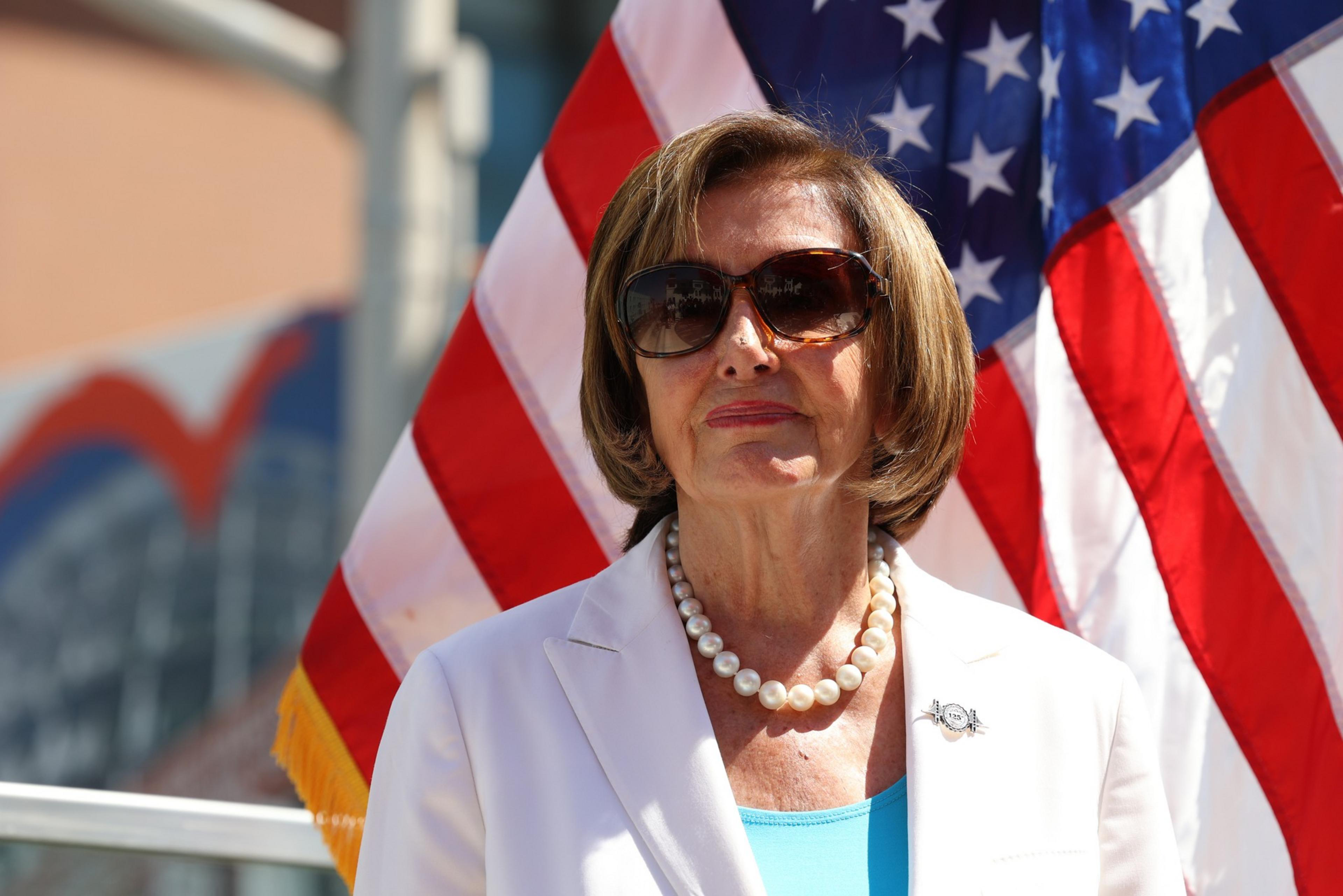 A woman wearing dark sunglasses, a white blazer, a blue top, and a pearl necklace stands in front of American flags.