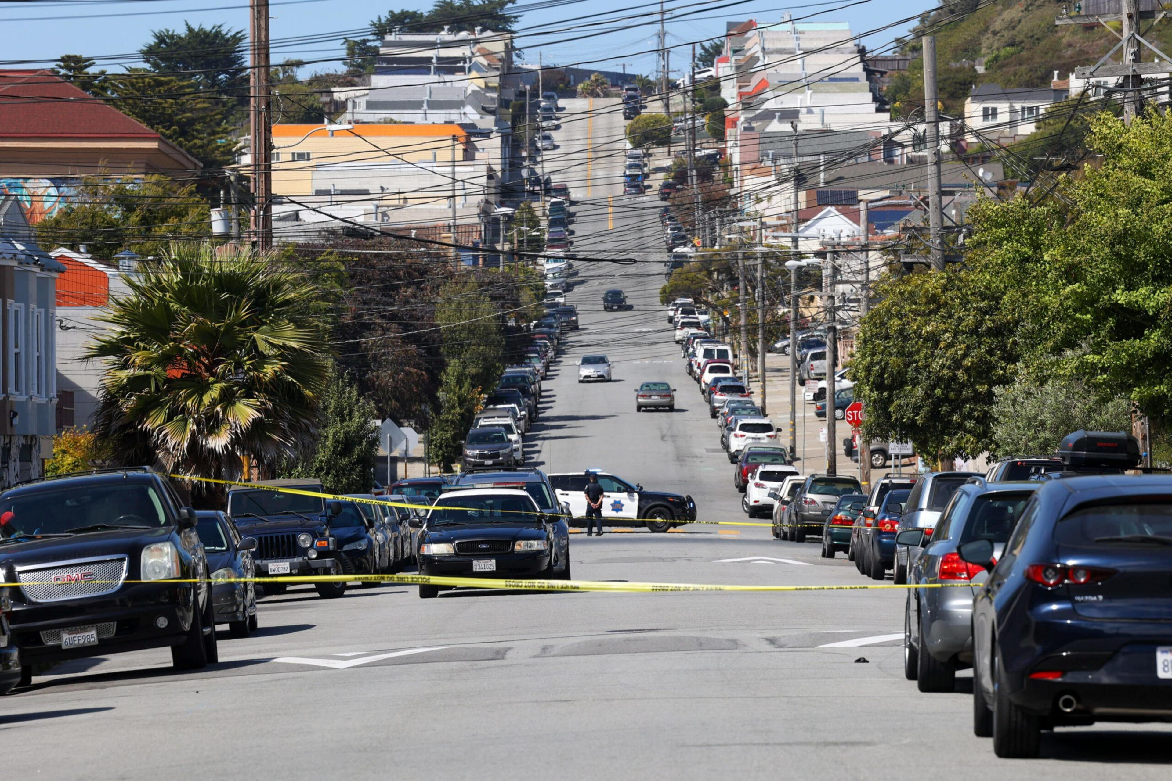 A long, steep street lined with parked cars on both sides is blocked by police tape and two police vehicles, with an officer standing in the middle.