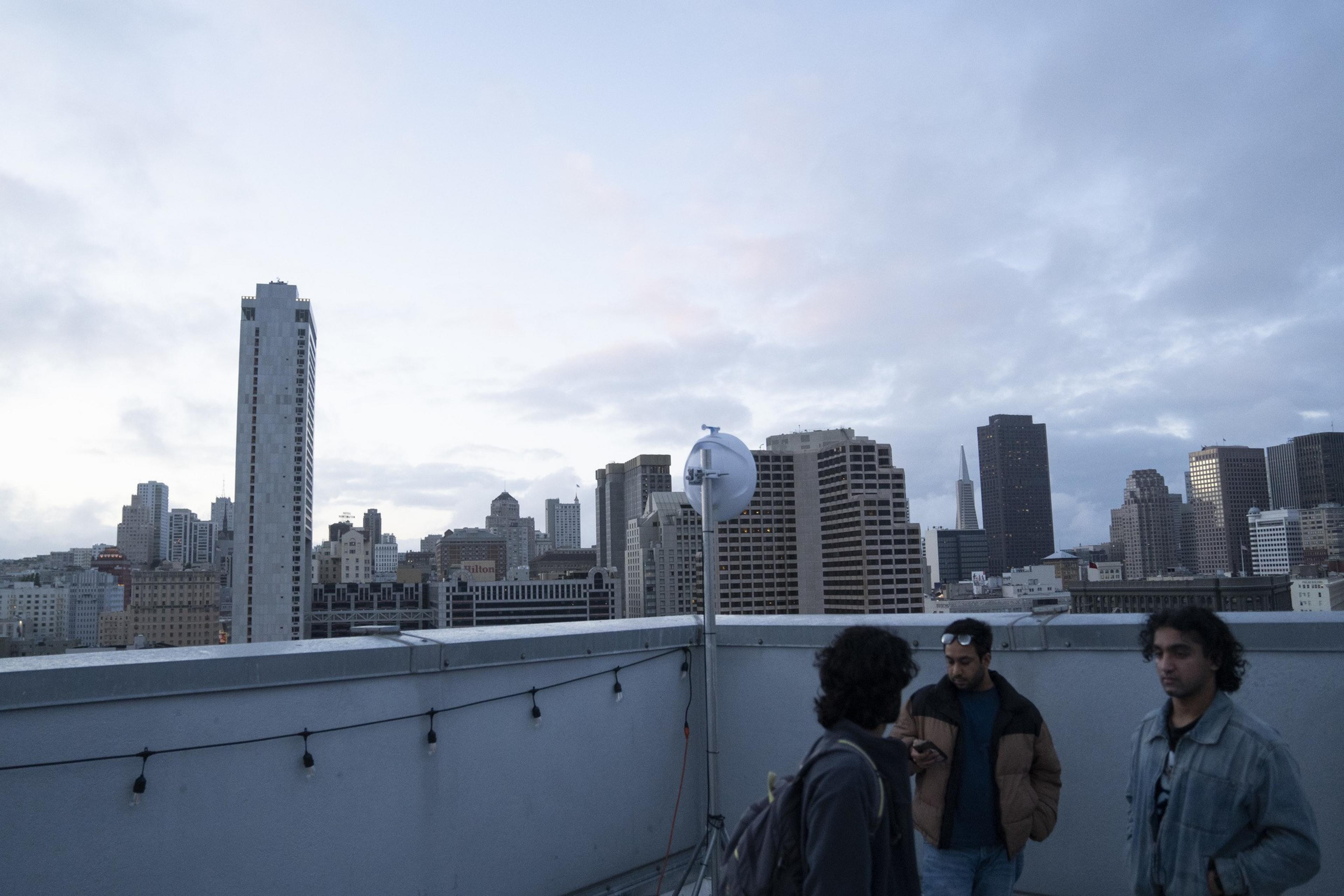 Three people are standing on a rooftop with a city skyline in the background. The sky is cloudy, and decorative lights and a satellite dish are visible.