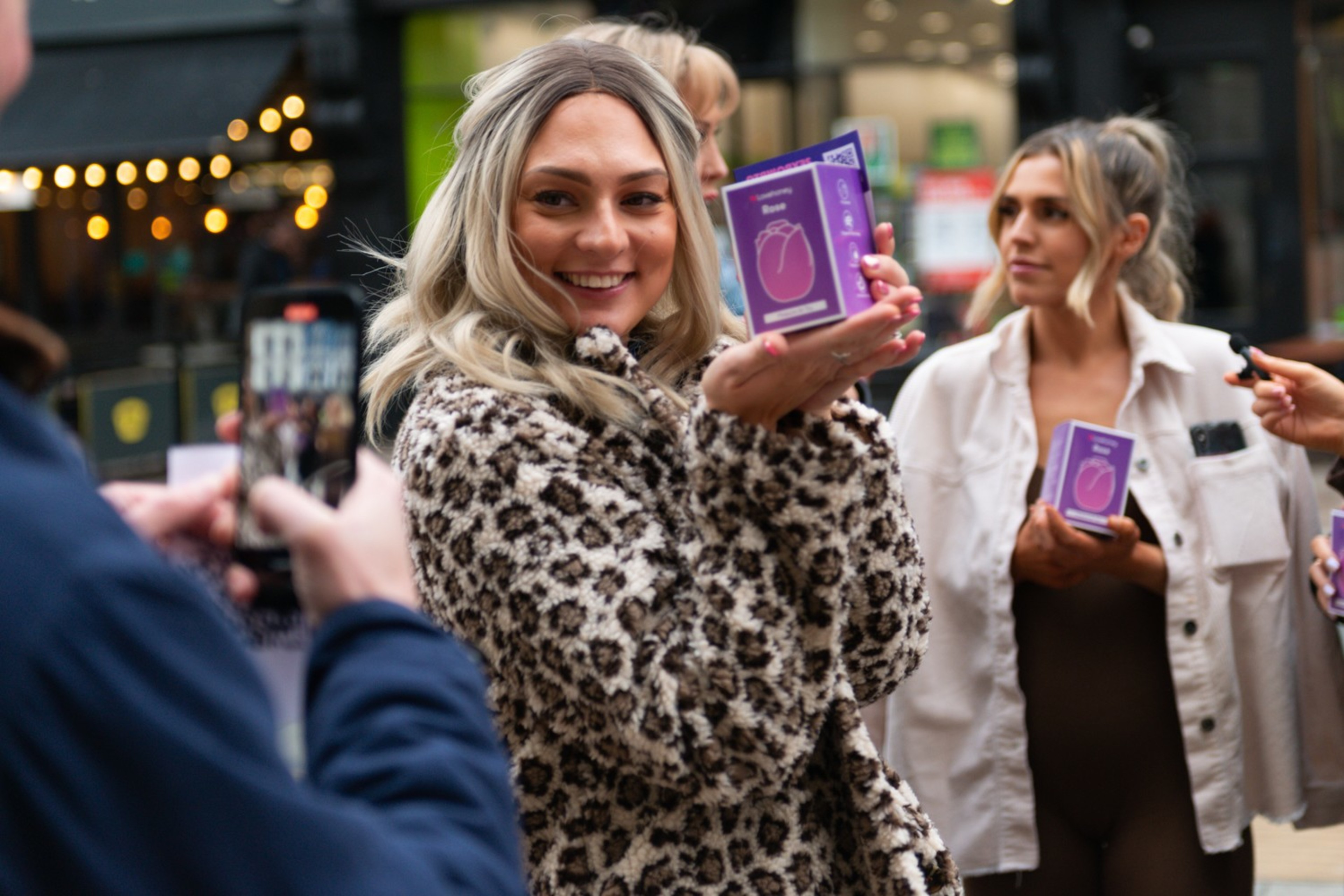 A woman in a leopard print coat smiles while holding a purple boxed product. Another person in white holds a similar box. A person nearby takes a photo.