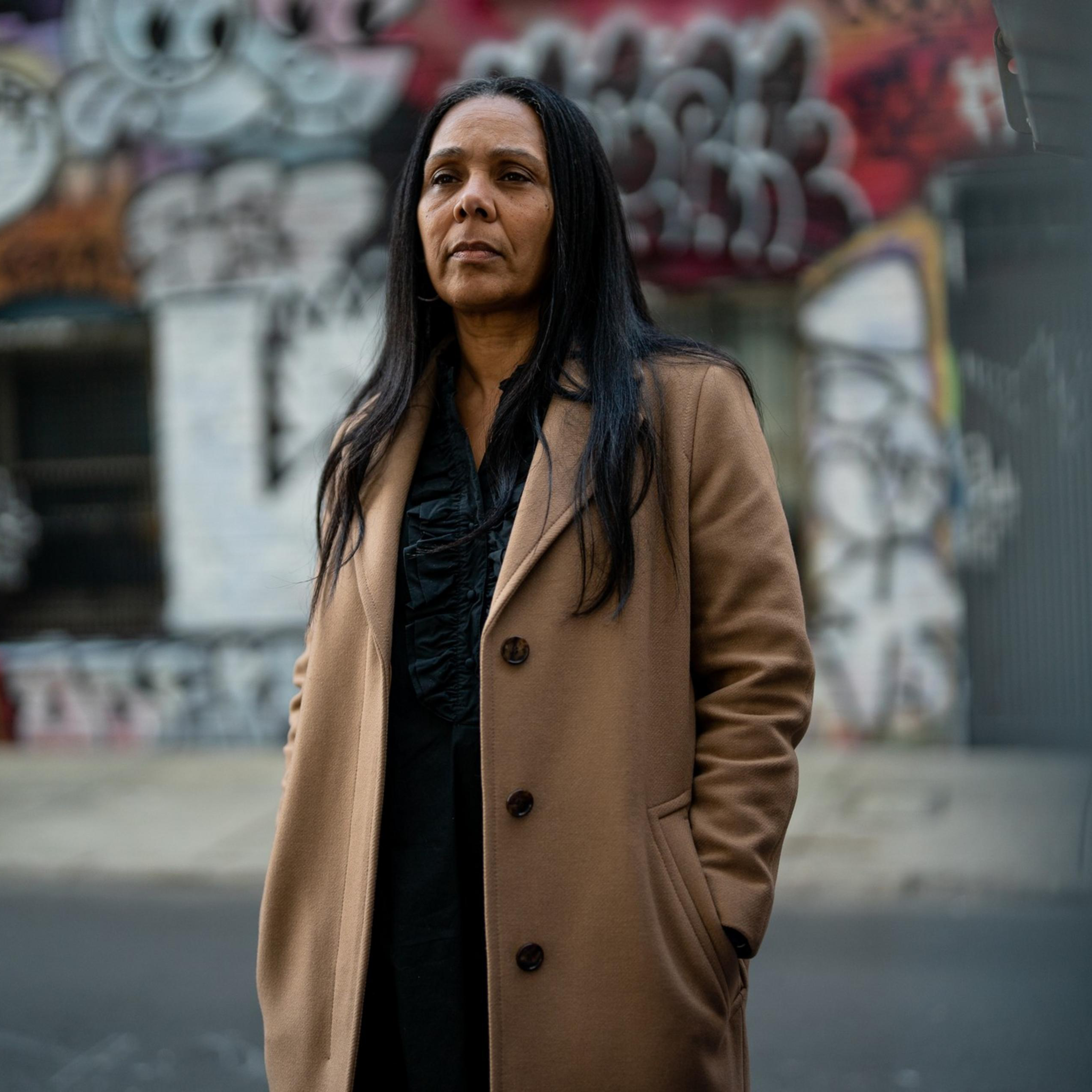 A woman in a brown coat stands in front of a wall covered with colorful graffiti. She has long dark hair and a thoughtful expression.