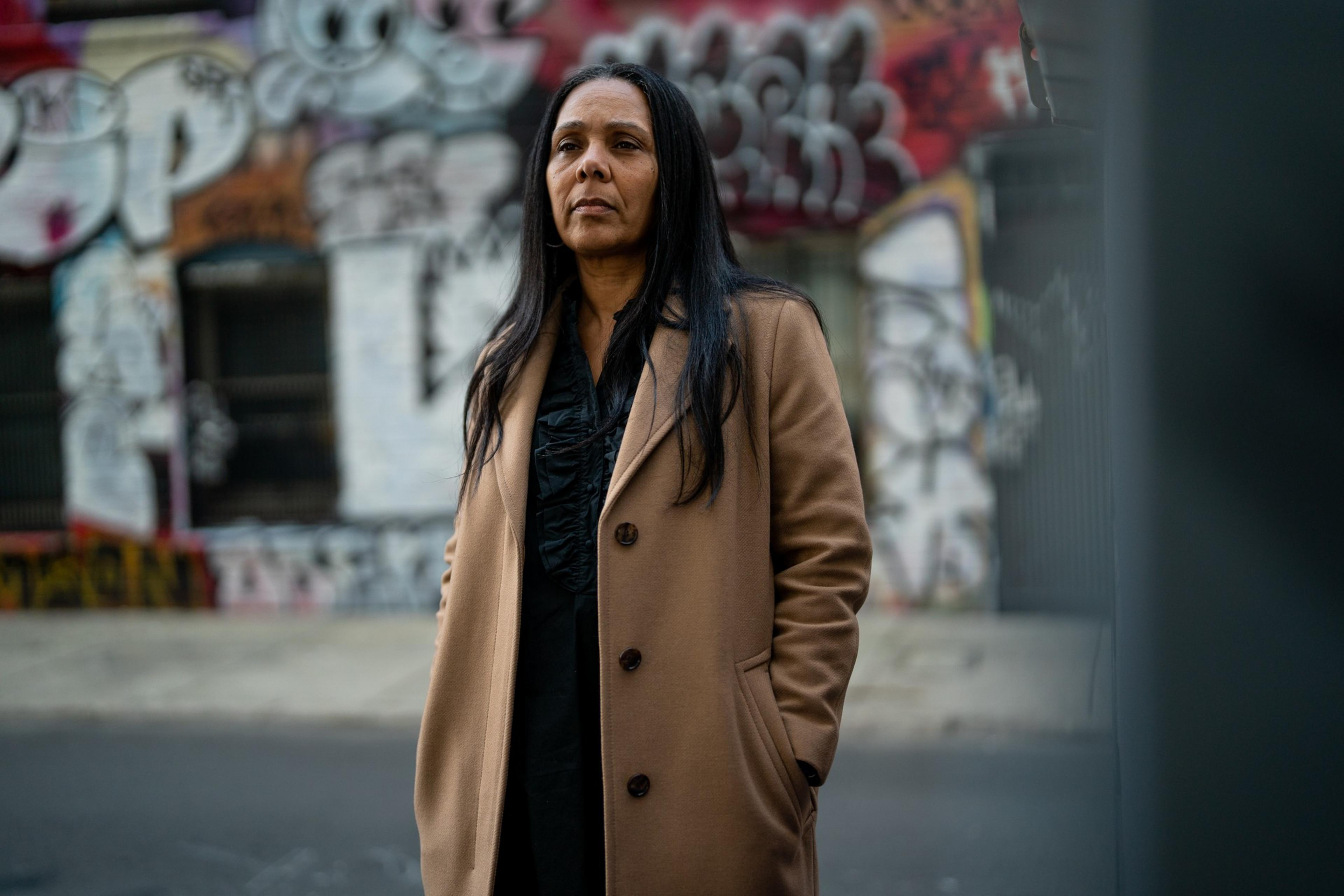 A woman in a brown coat stands in front of a wall covered with colorful graffiti. She has long dark hair and a thoughtful expression.
