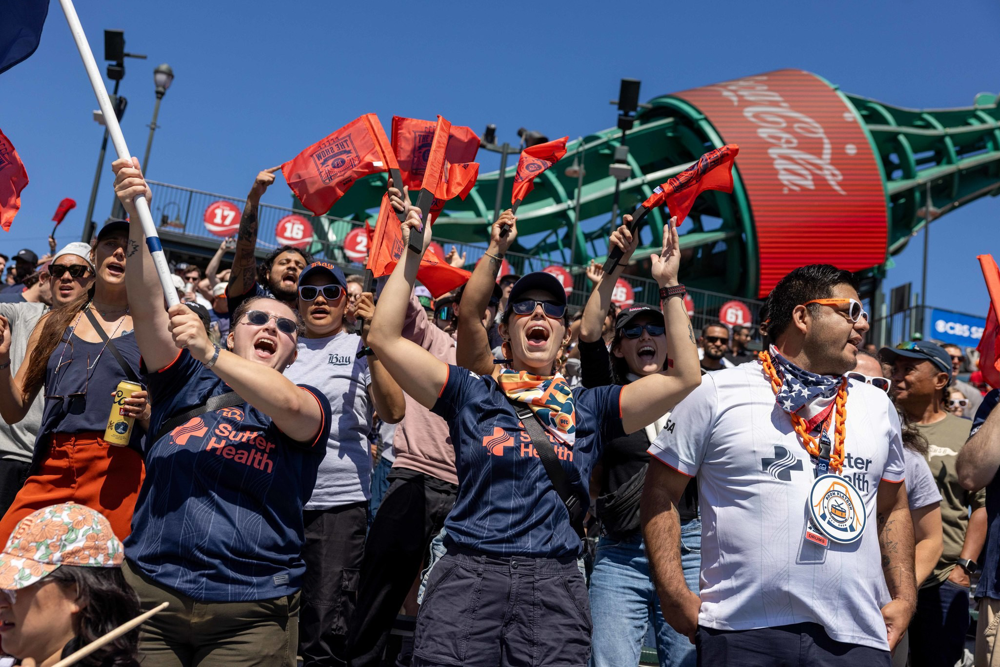 Fans wearing Sacramento Republic FC shirts cheer and wave orange flags in front of a large Coca-Cola bottle sculpture at a stadium.