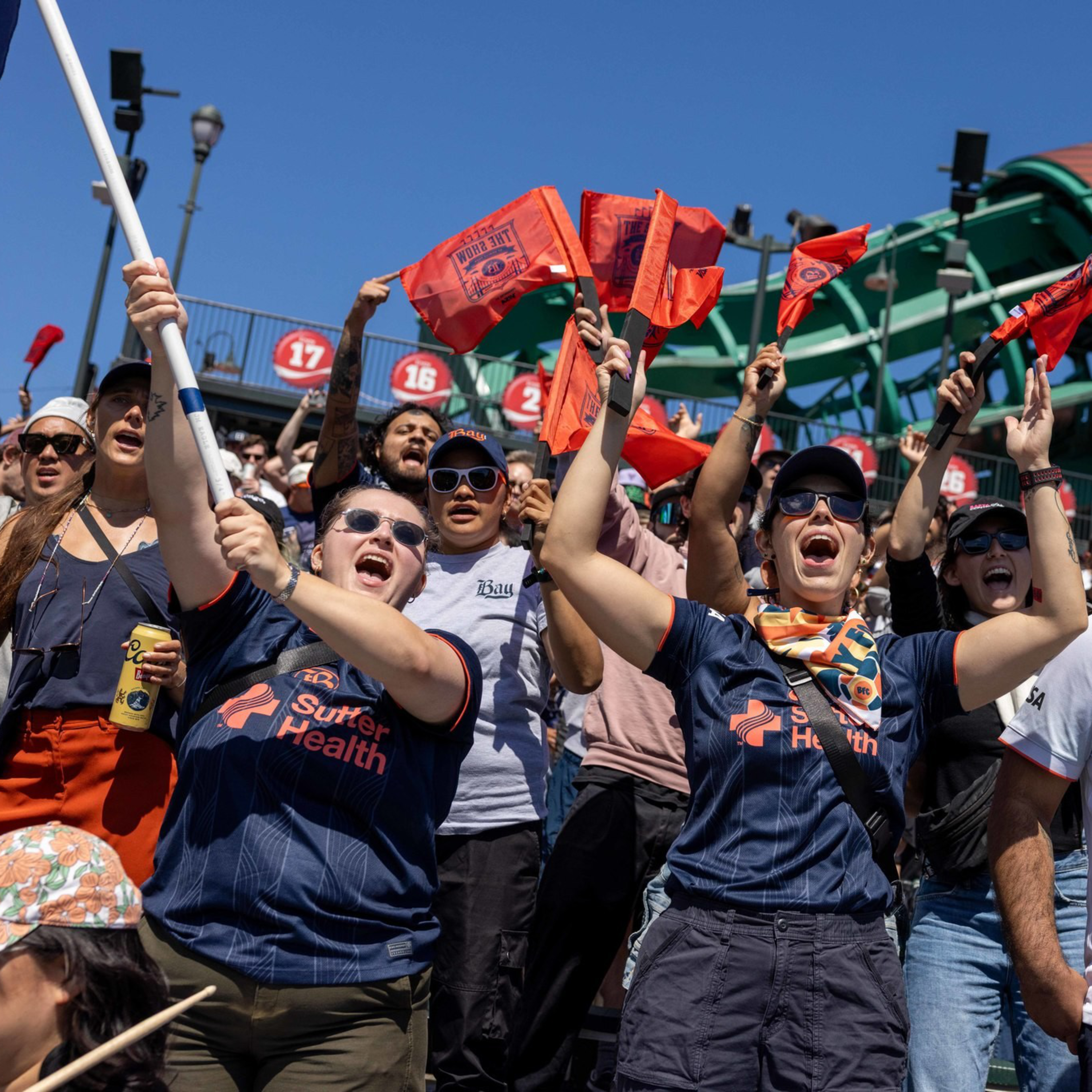 Fans wearing Sacramento Republic FC shirts cheer and wave orange flags in front of a large Coca-Cola bottle sculpture at a stadium.