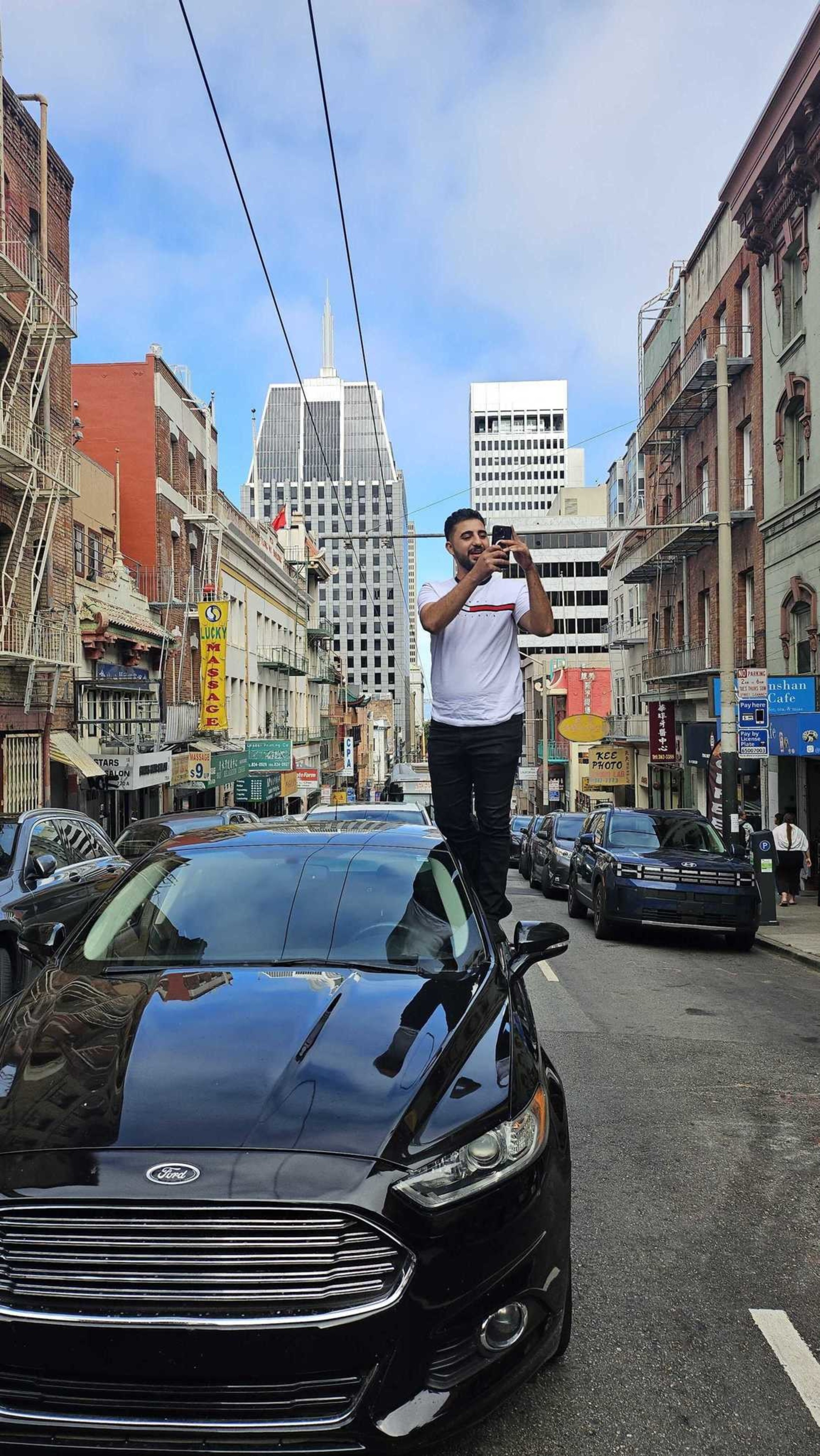 A man stands on the roof of a black Ford car, taking a photo with his phone in a busy urban street lined with buildings and parked cars.