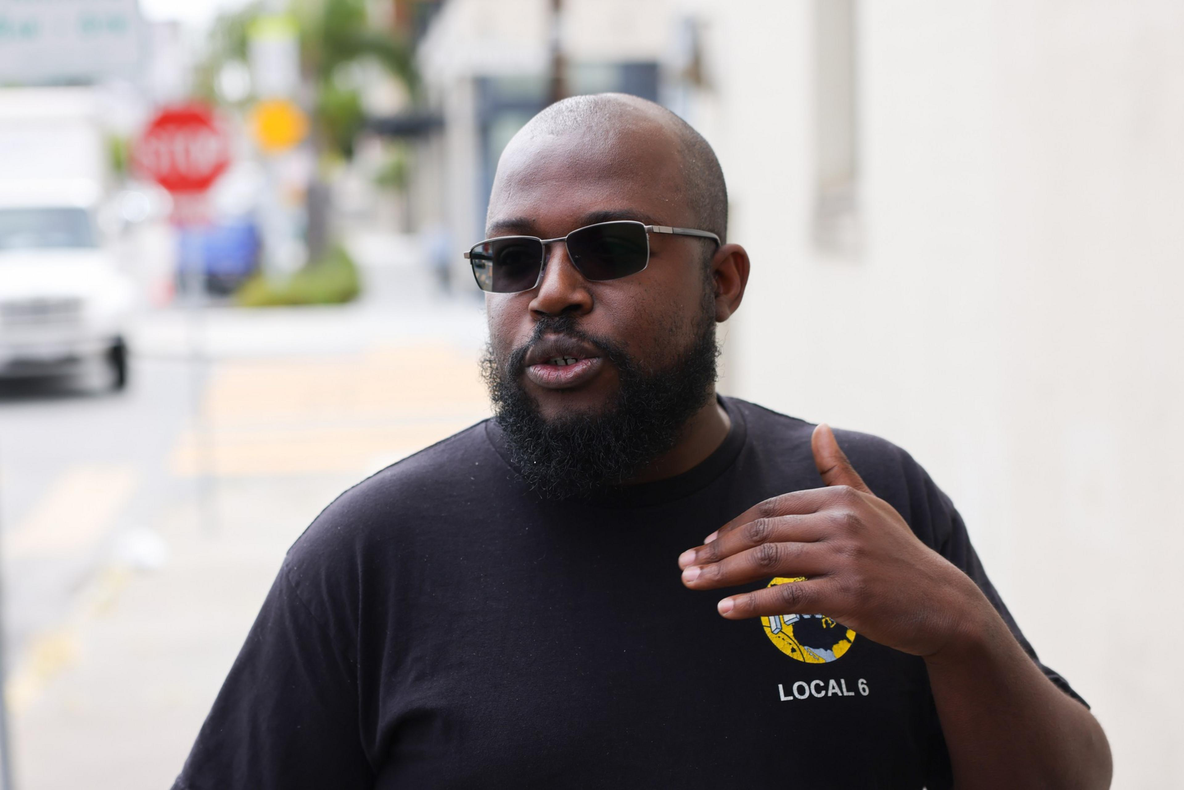 A man with a beard and sunglasses wears a black shirt with a yellow "LOCAL 6" logo while gesturing with his right hand.