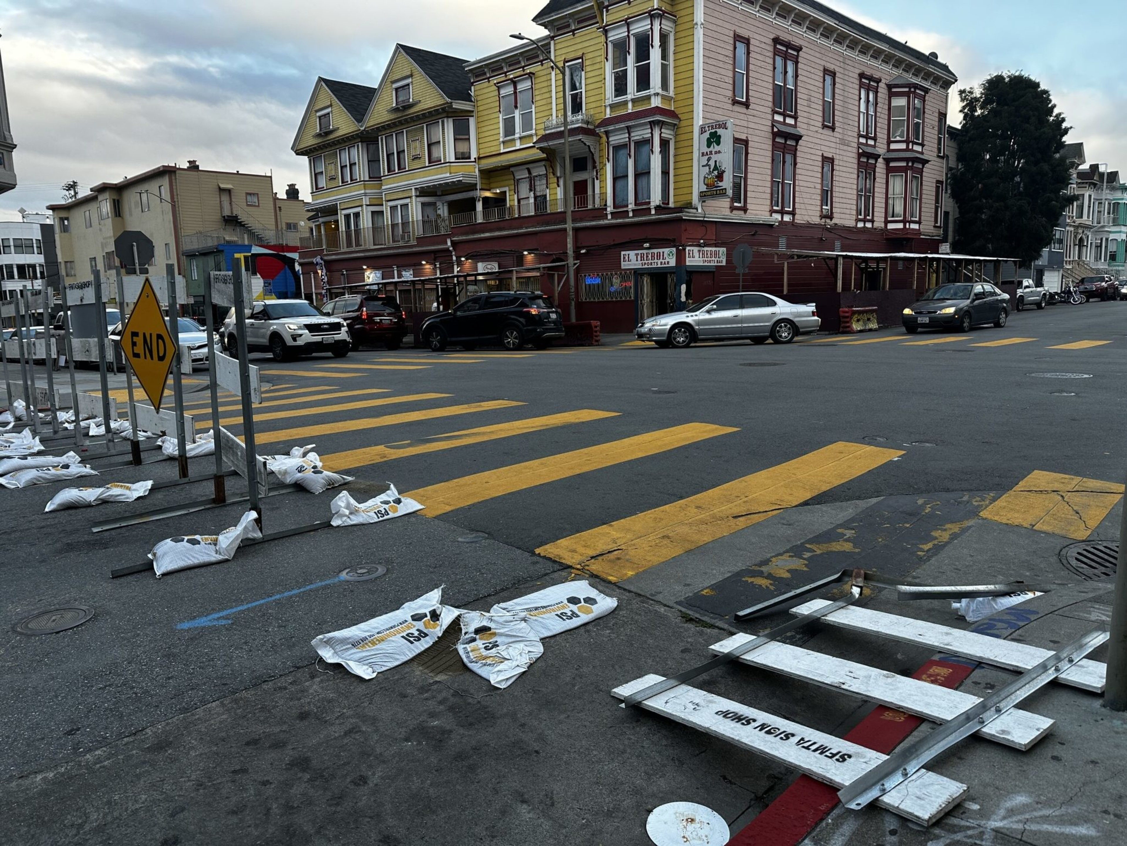A crosswalk with bold yellow stripes spans the street, bordered by old buildings and cars, while sandbags and fallen signs clutter the sidewalk.