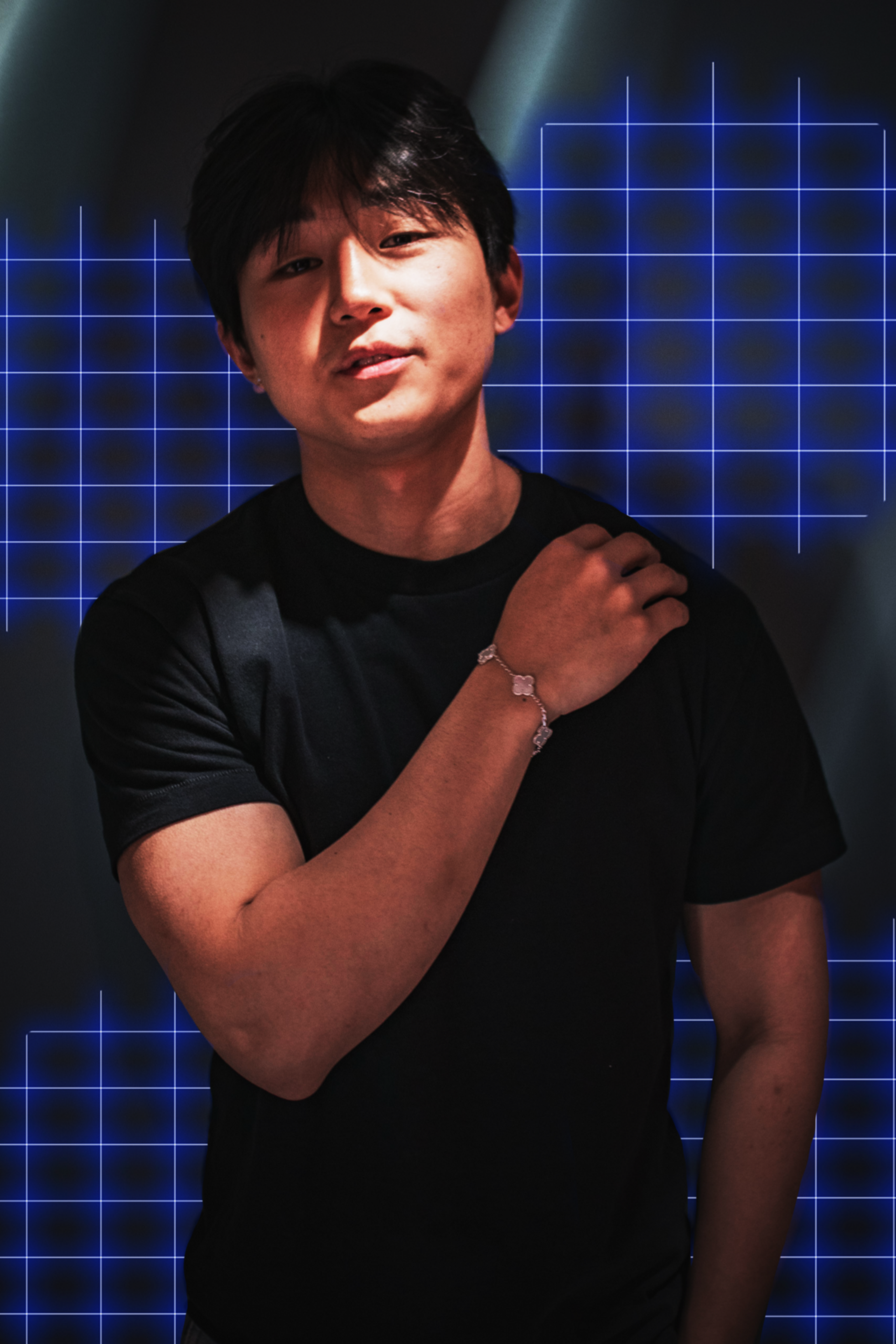 A young man with black hair wears a black t-shirt and a silver bracelet, with a blue grid pattern glowing softly in the dark background.