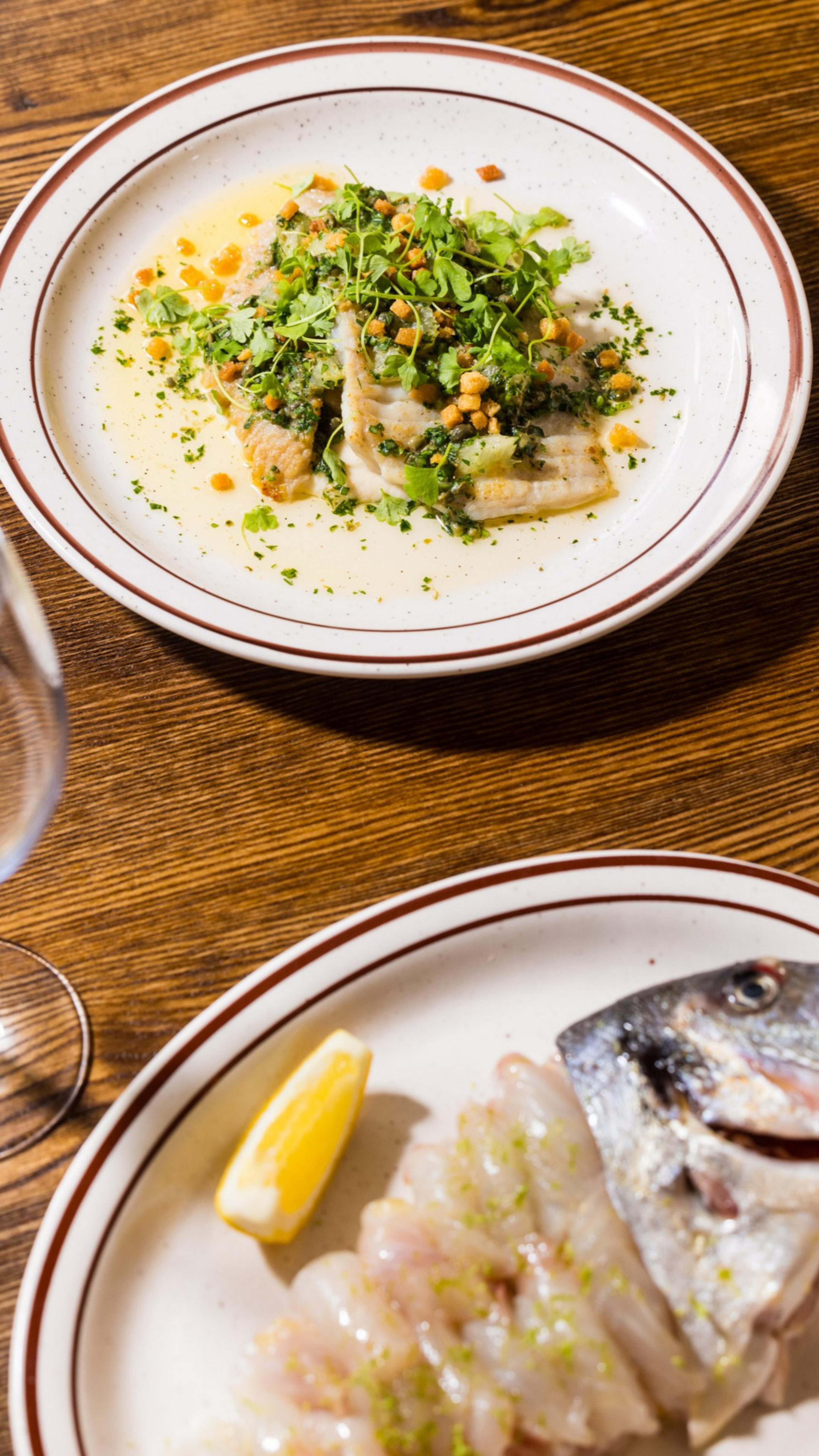 Two dishes are on a wooden table. The top plate has fish with herbs and croutons, while the bottom plate shows a sliced fish with a lemon wedge. A glass is partially visible.