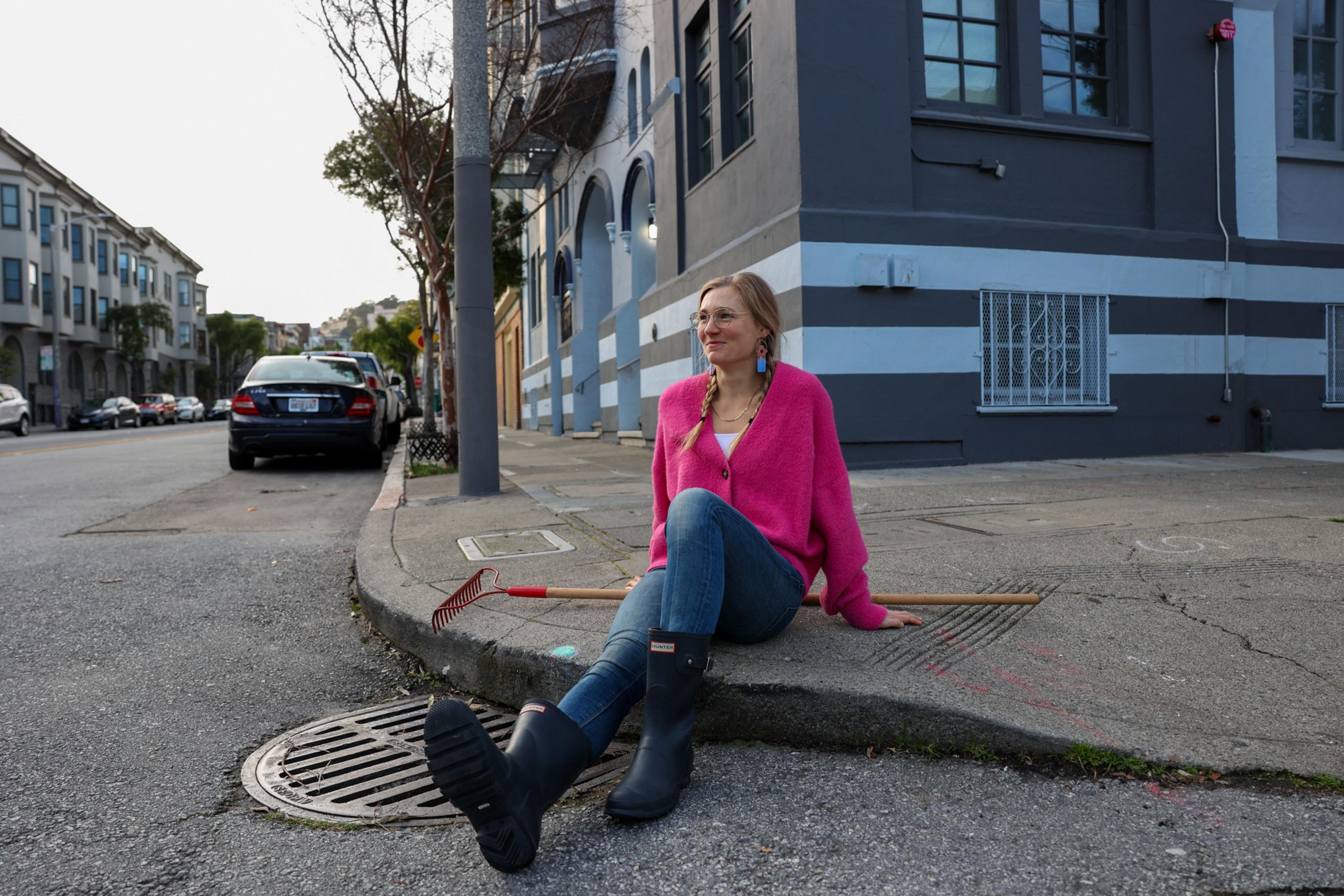 A woman in a bright pink sweater, jeans, and black rain boots sits on a curb, smiling, with two garden rakes beside her on an urban street corner.