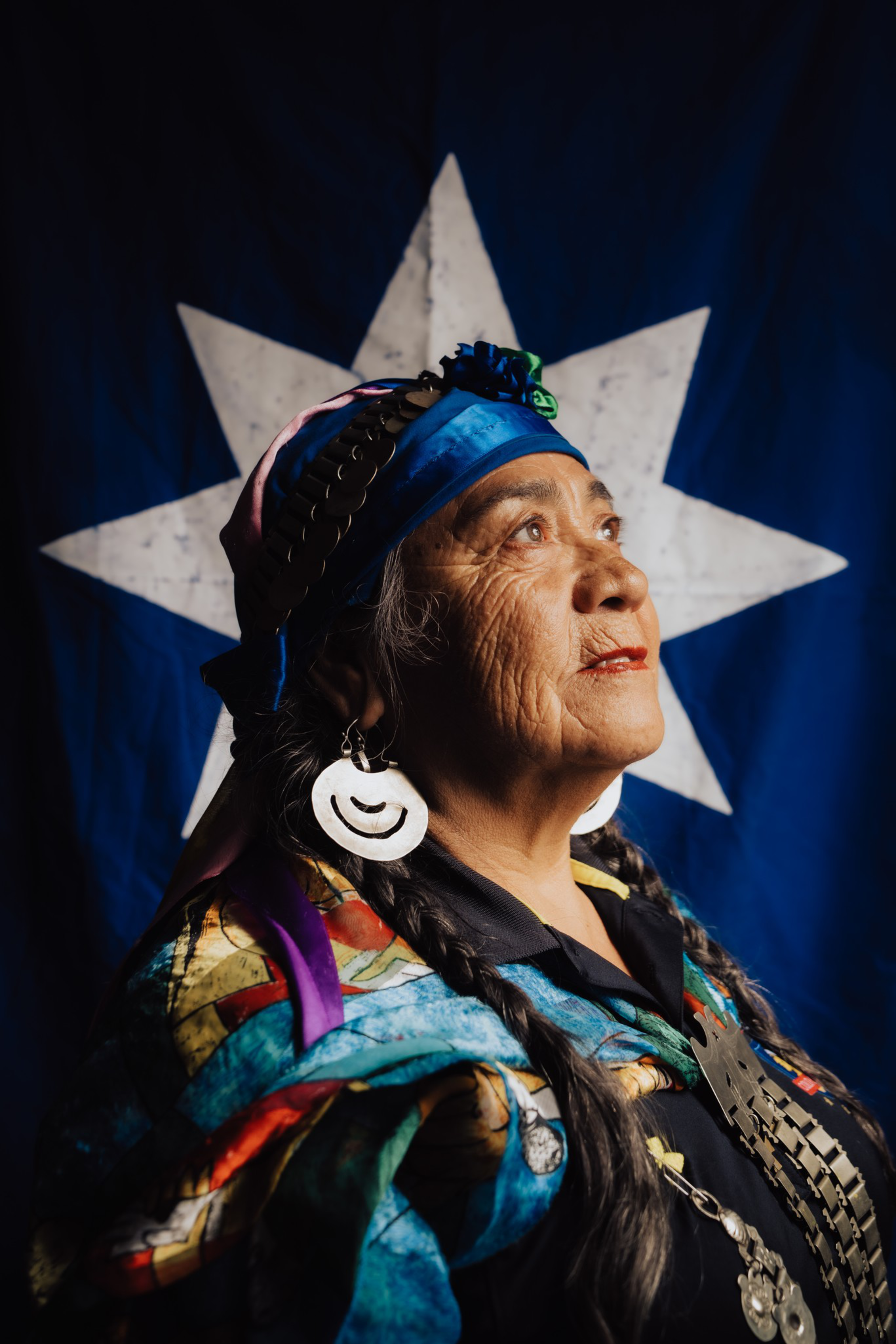 An elder woman wearing colorful traditional clothing and large round earrings looks upward, standing against a dark blue background with a large white star.