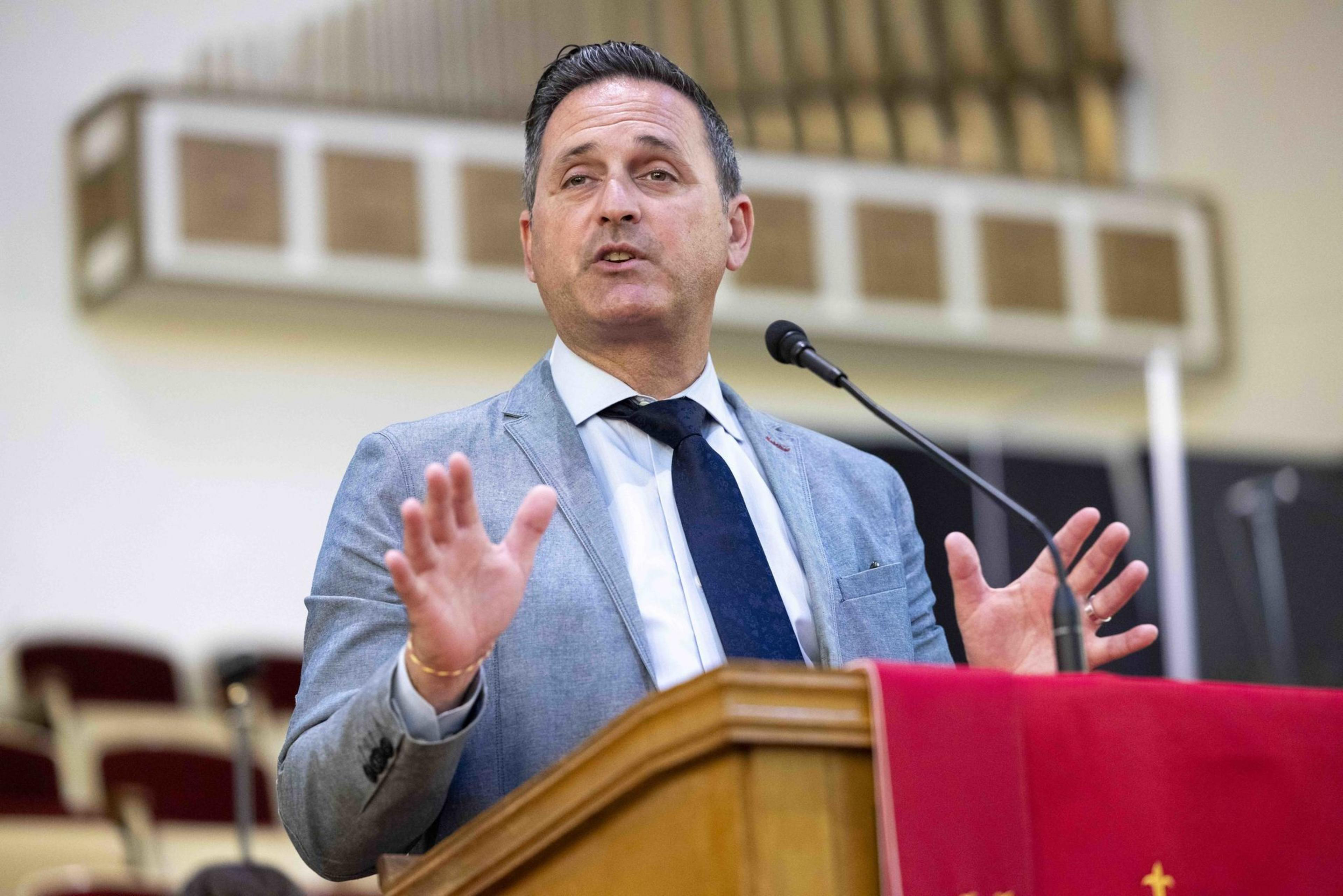 A man in a blue suit and tie is speaking at a podium with a red cloth and microphone, gesturing with his hands. Organ pipes are visible in the background.