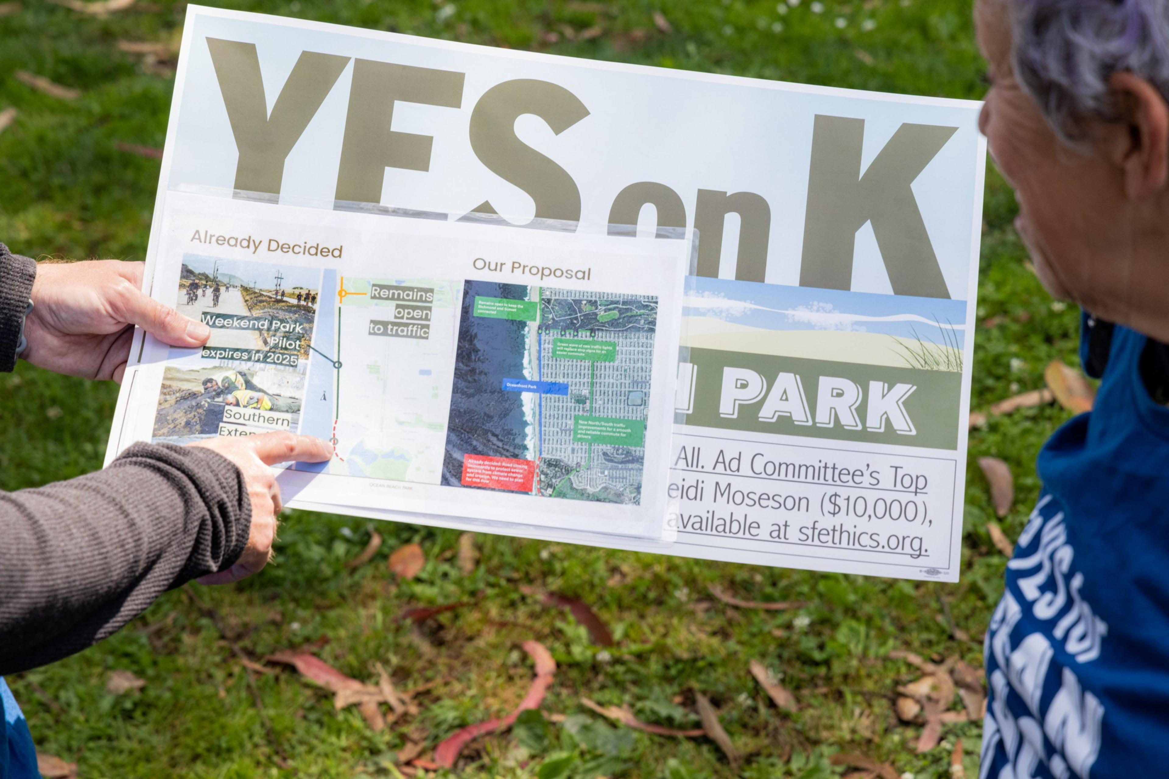 A person holds a sign that says &quot;Yes on K.&quot; It includes a map and text about a park proposal and traffic plans. Two people are discussing it outdoors.