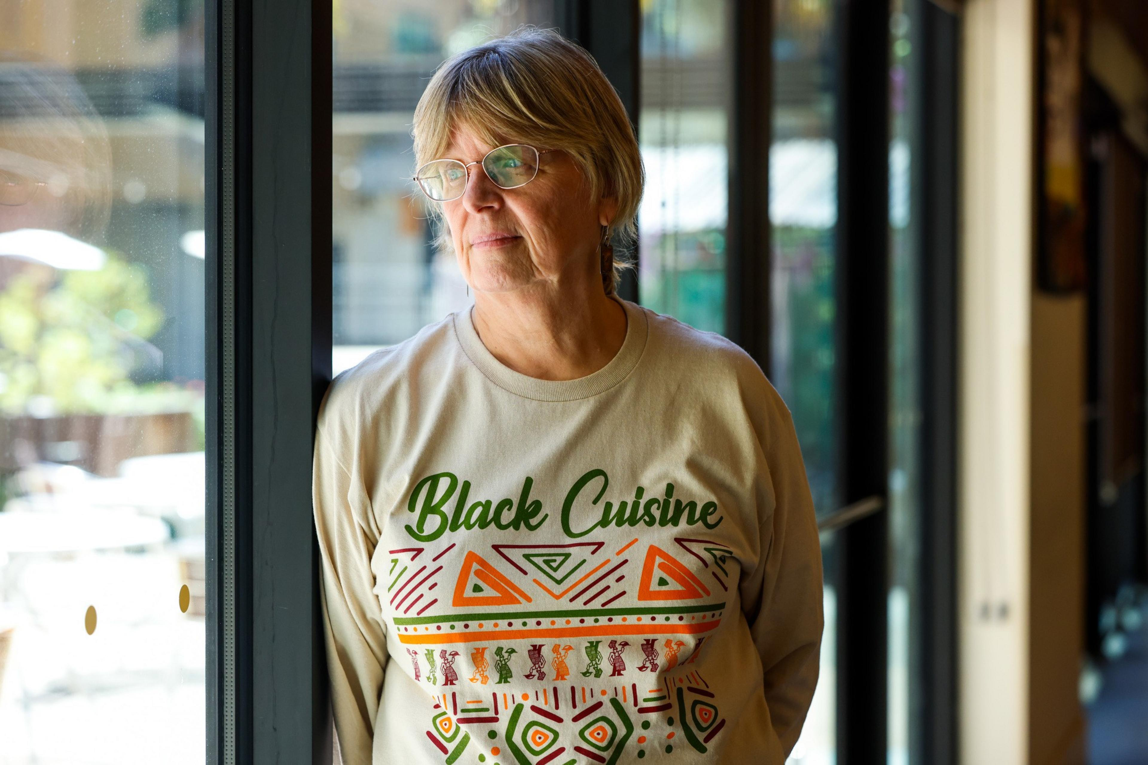 A person with short hair and glasses stands near a window, wearing a shirt with &quot;Black Cuisine&quot; and colorful geometric patterns.