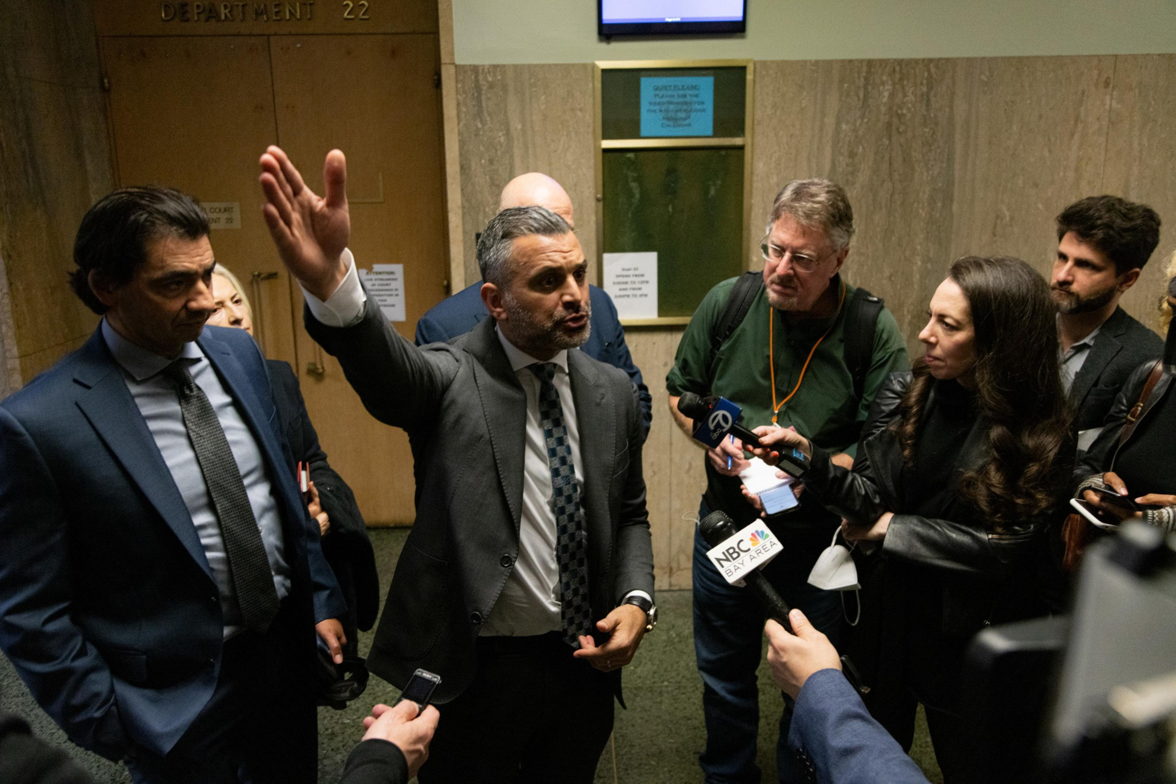 A group of people, including a man gesturing, stand outside a courtroom surrounded by reporters with microphones.