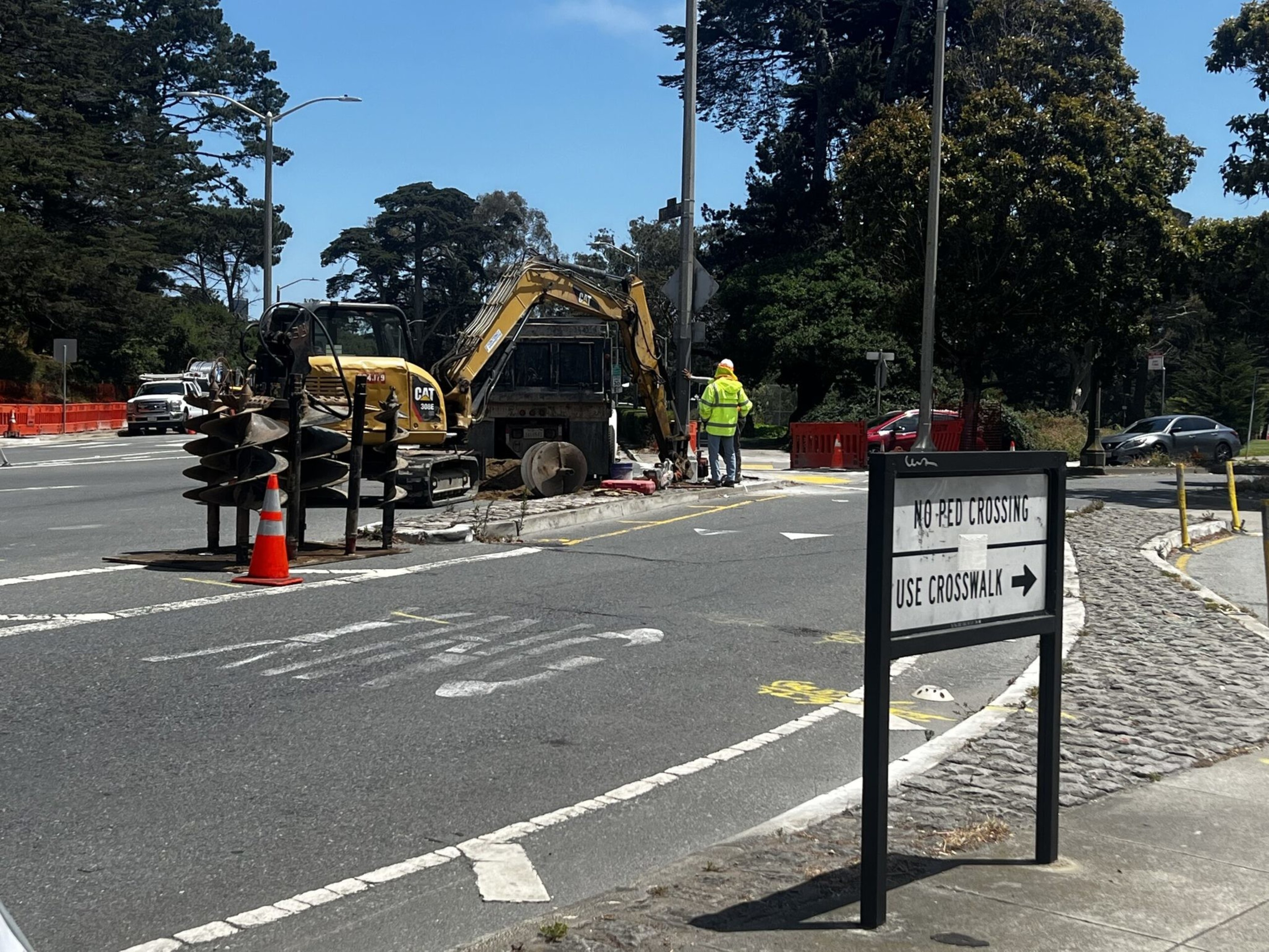 Road construction with an excavator, workers in safety gear, and a sign directing pedestrians to use the crosswalk instead of crossing here.
