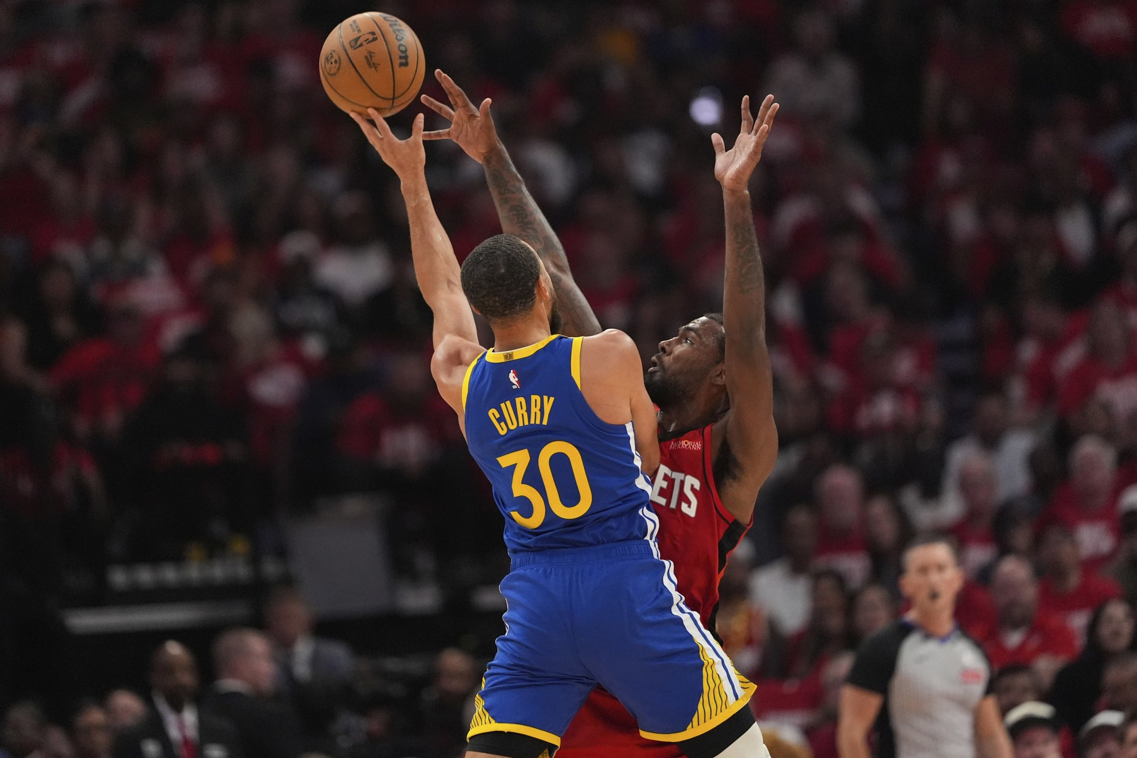 A basketball player in a blue jersey jumps to shoot, while another in a red jersey defends with arms raised. The crowd watches intensely from the background.