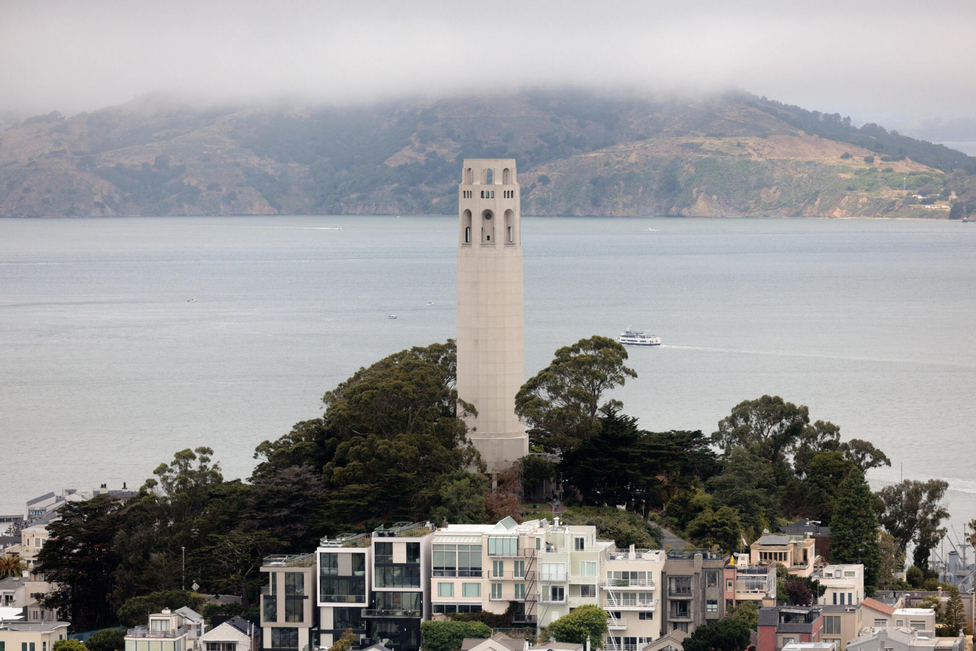 A tall white tower rises above a cluster of trees on a hill, with modern buildings in the foreground and mist-covered hills across calm water in the background.