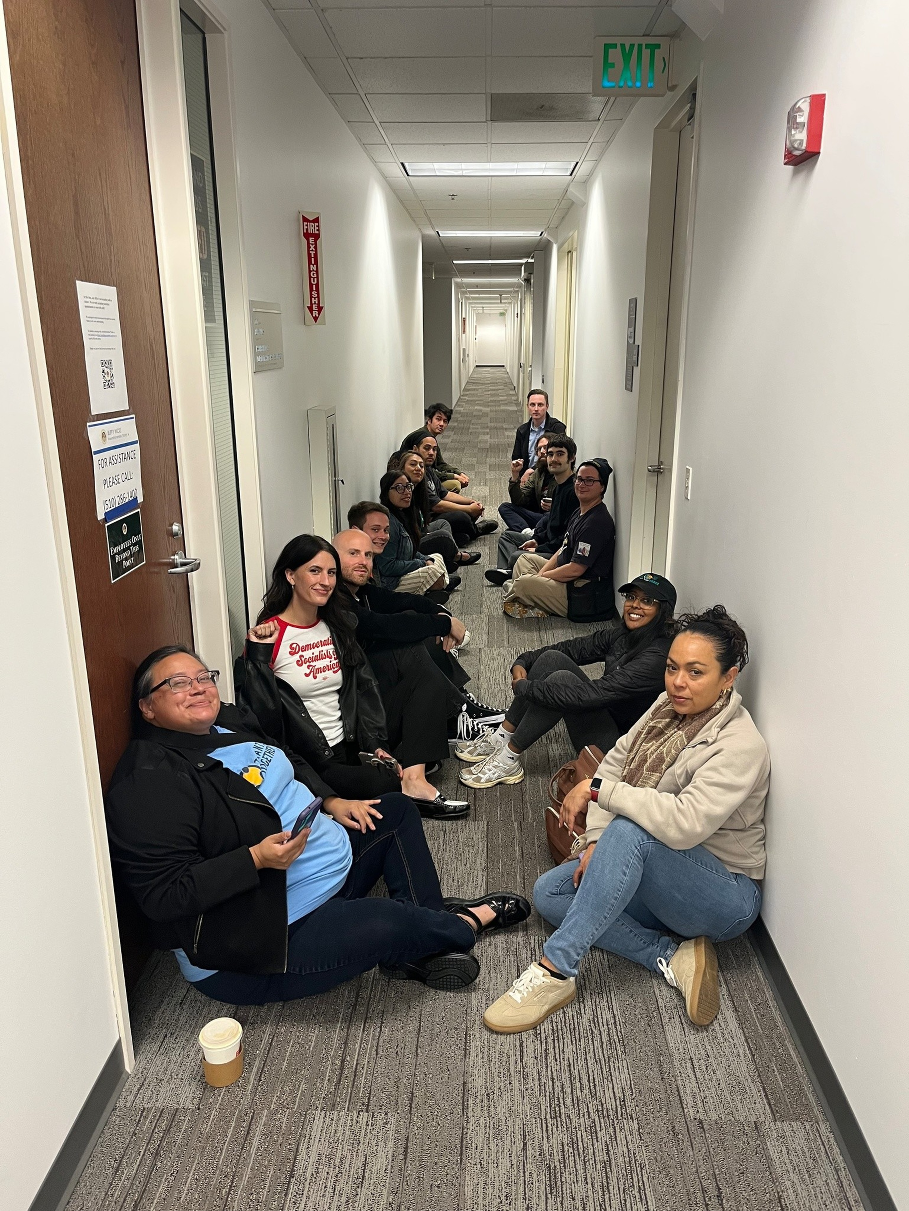 A group of 14 people sitting casually on the floor in a narrow hallway, smiling and facing the camera, with white walls and carpeted floor.