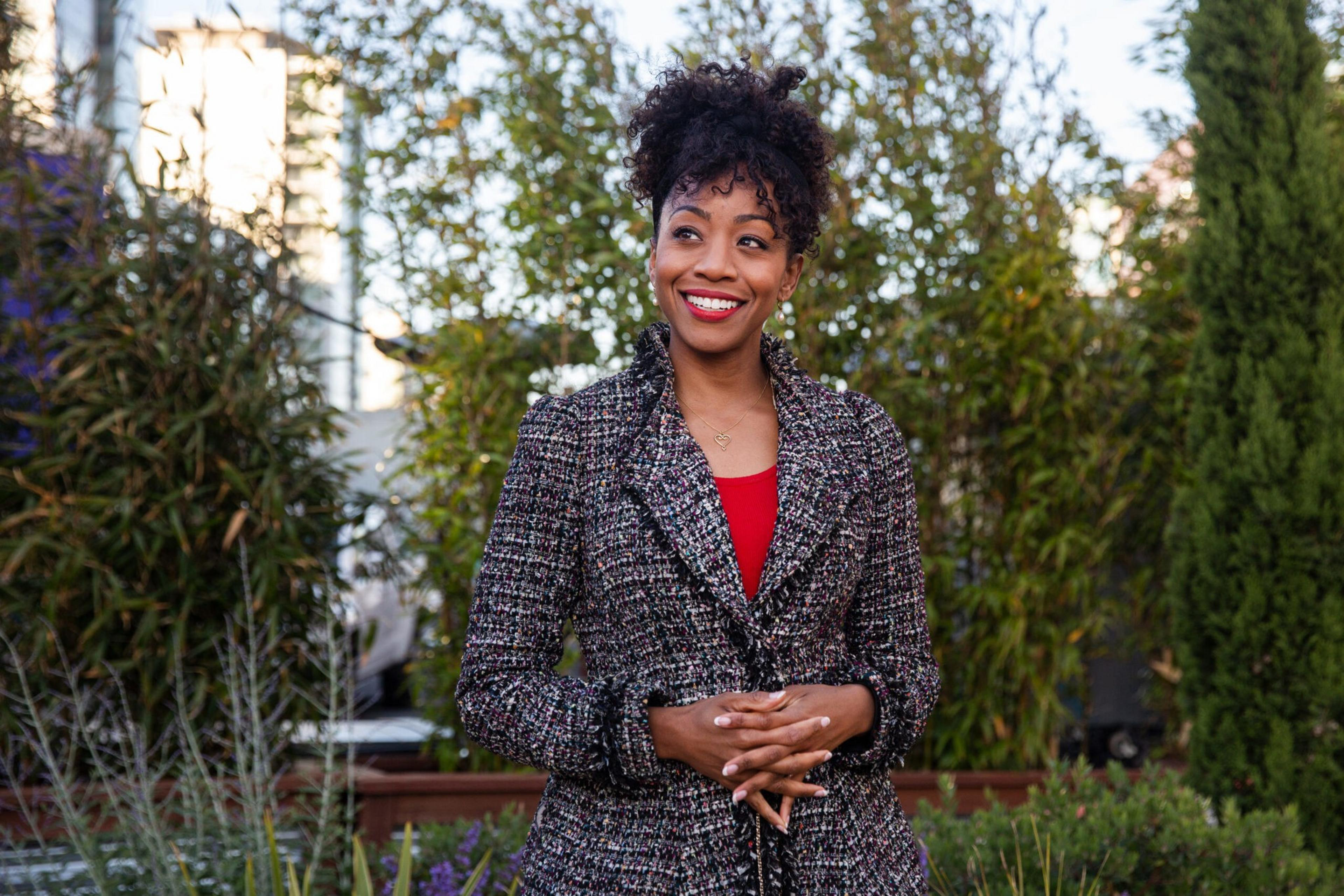 A woman with curly hair smiles warmly, wearing a textured jacket over a red top, standing in a garden with green trees and plants around her.