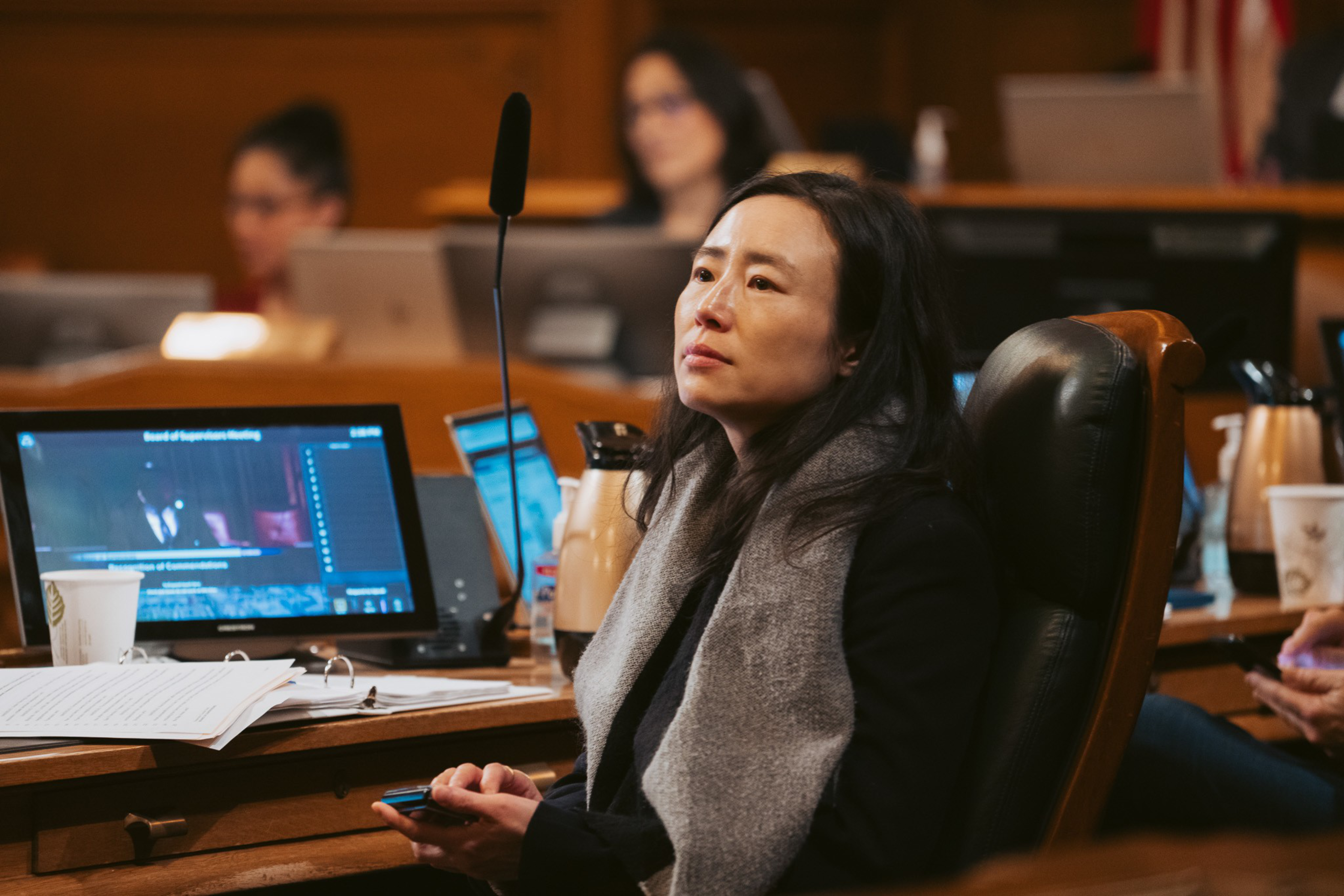 A woman with long dark hair and a gray scarf sits in a leather chair, looking thoughtfully to the side in a wood-paneled room with computer screens and papers.