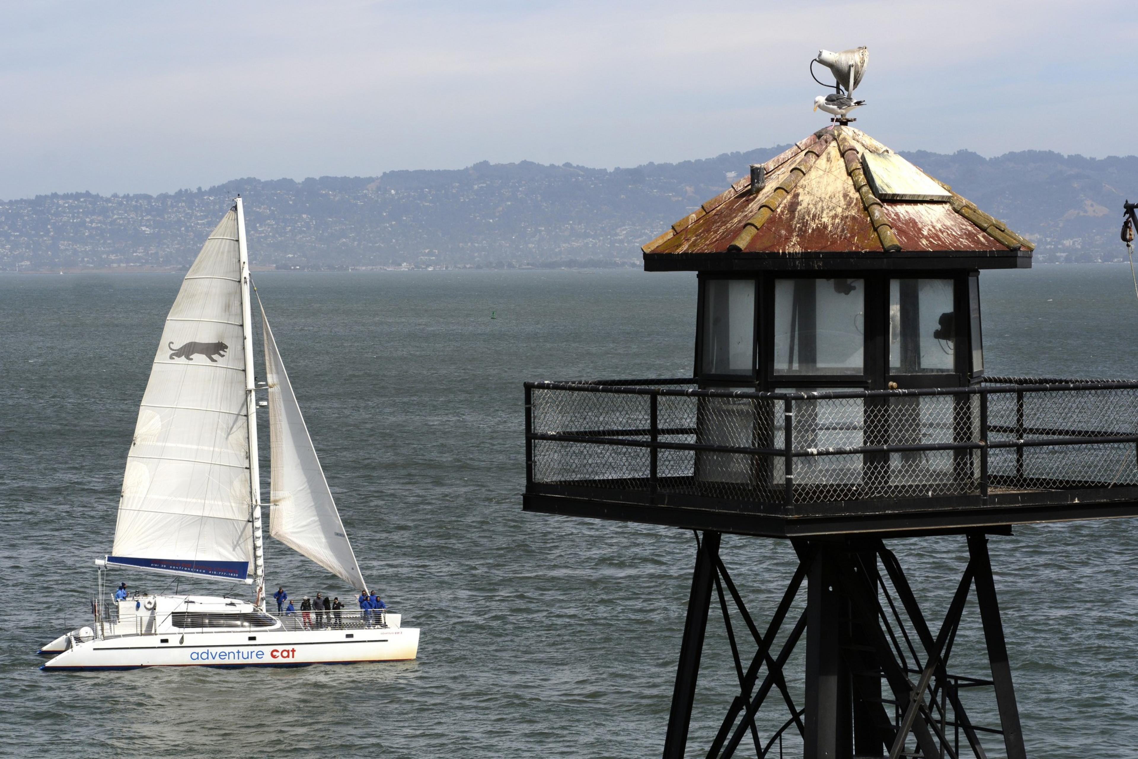 A sailboat named &quot;Adventure Cat&quot; glides on water near a black metal tower. The background shows distant hills under a lightly clouded sky.