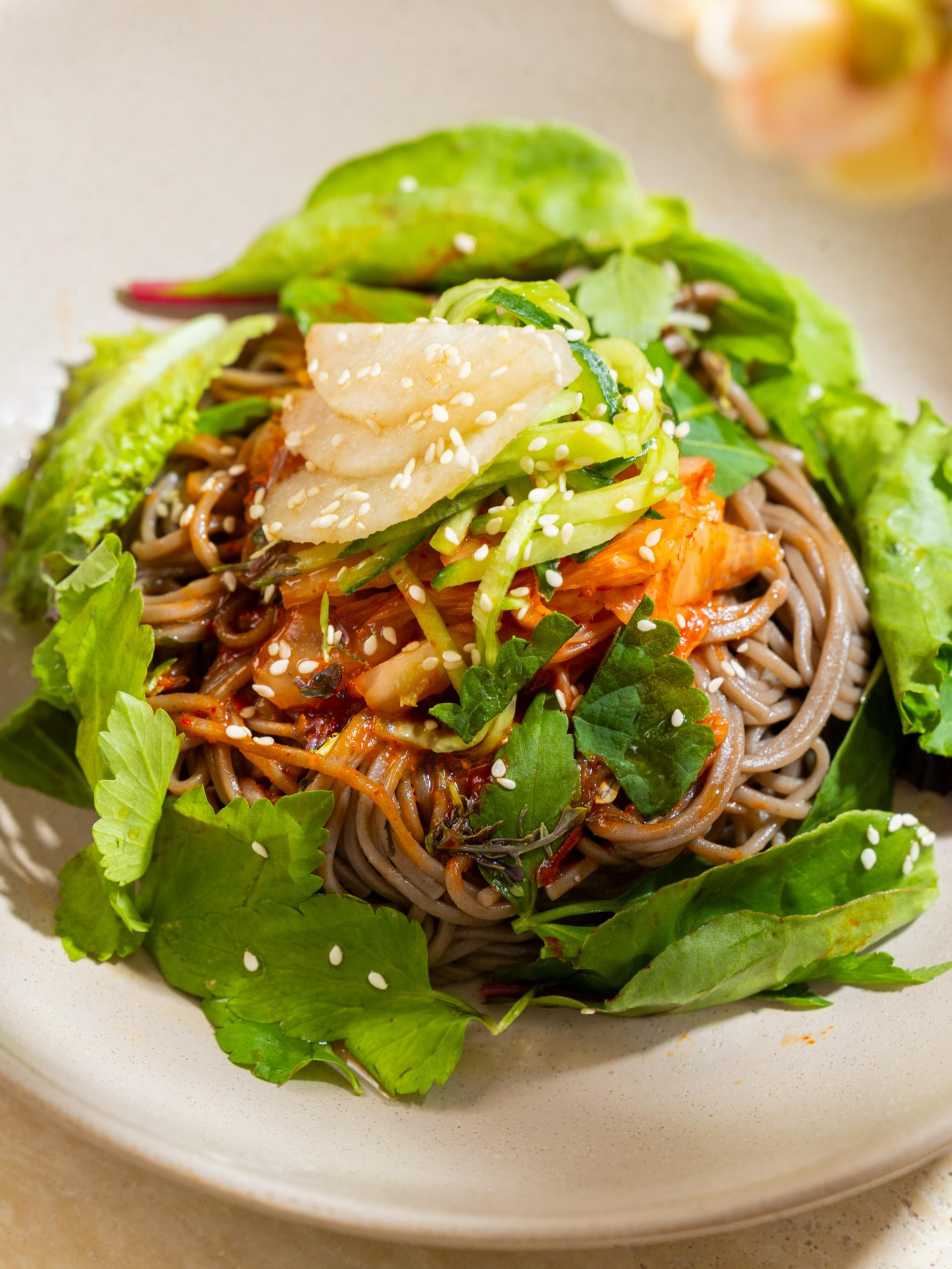 Brown soba noodles topped with sliced cucumbers, radish, kimchi, and sesame seeds, garnished with fresh green leafy herbs on a beige plate.