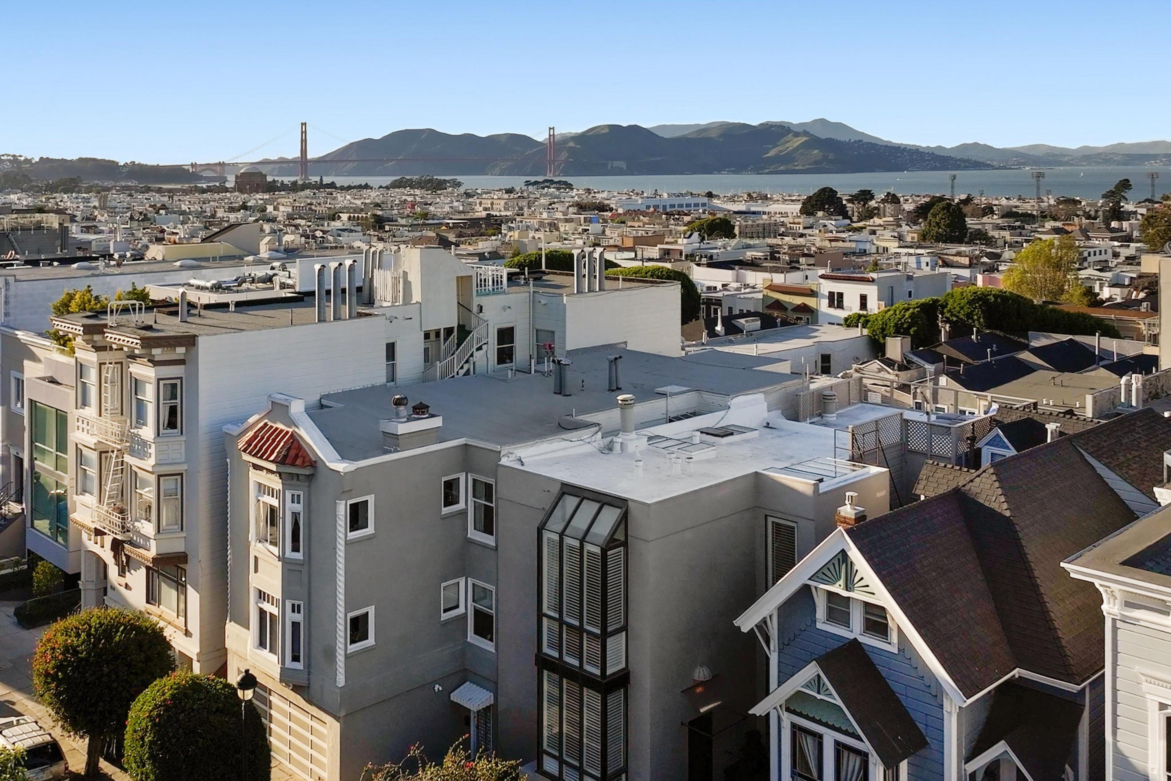 The image shows a cityscape with tightly packed buildings, trees, and the Golden Gate Bridge in the background, under a clear blue sky.